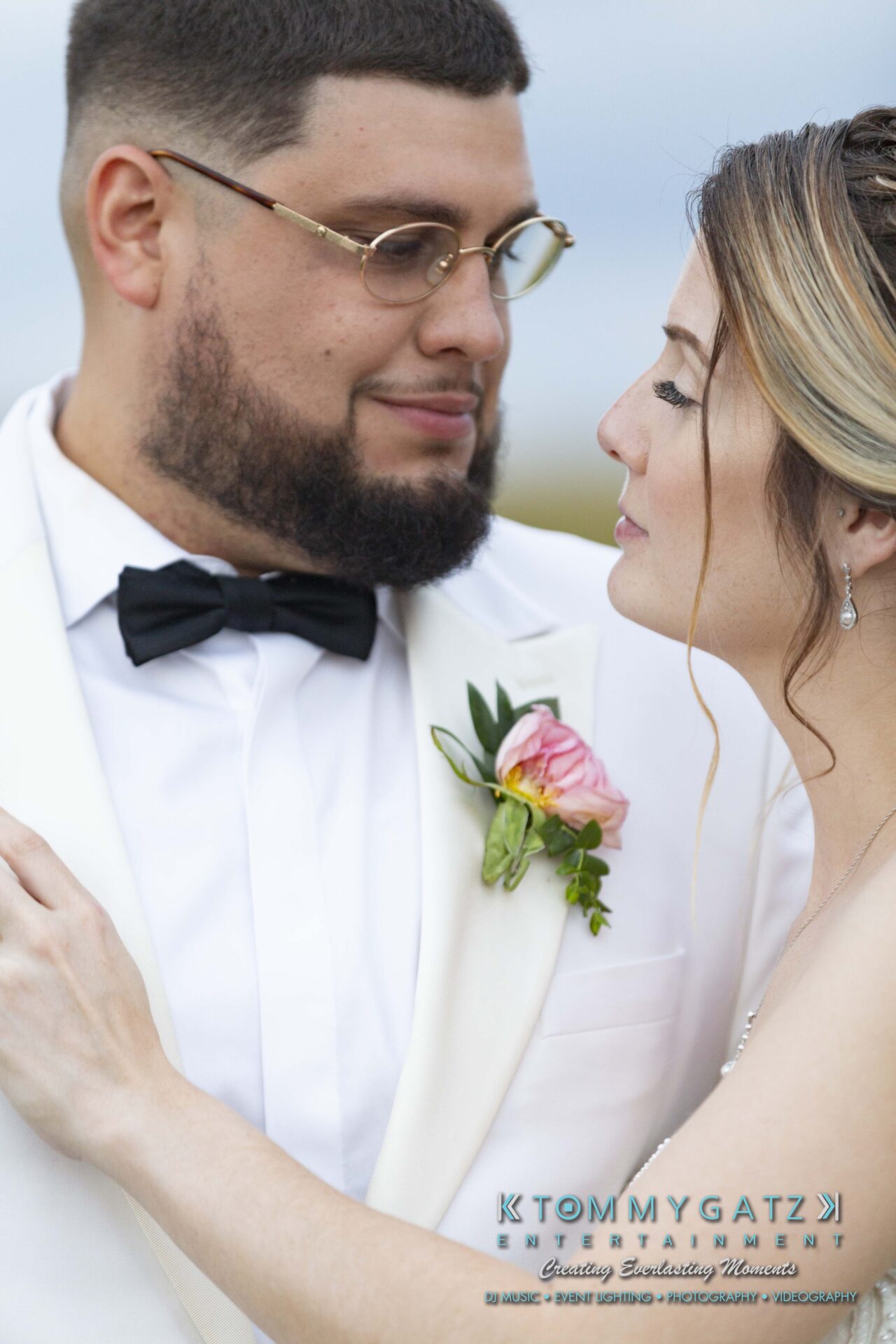 Kari-Jose-062318-FORMALS-Low-Res-WM-418 - TOMMY GATZ ENTERTAINMENT A couple in wedding attire gazes at each other lovingly. The groom wears a white suit and bow tie, while the bride has an updo hairstyle and a flower on her dress.