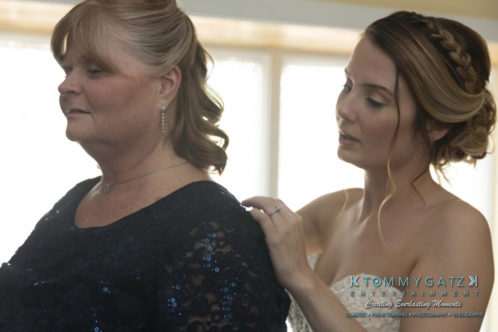 Kari-Jose-062318-PREP-Low-Res-WM-45 - TOMMY GATZ ENTERTAINMENT A bride assists a woman in a dark dress, adjusting the shoulder area. They are indoors, with natural light shining through the window behind them.