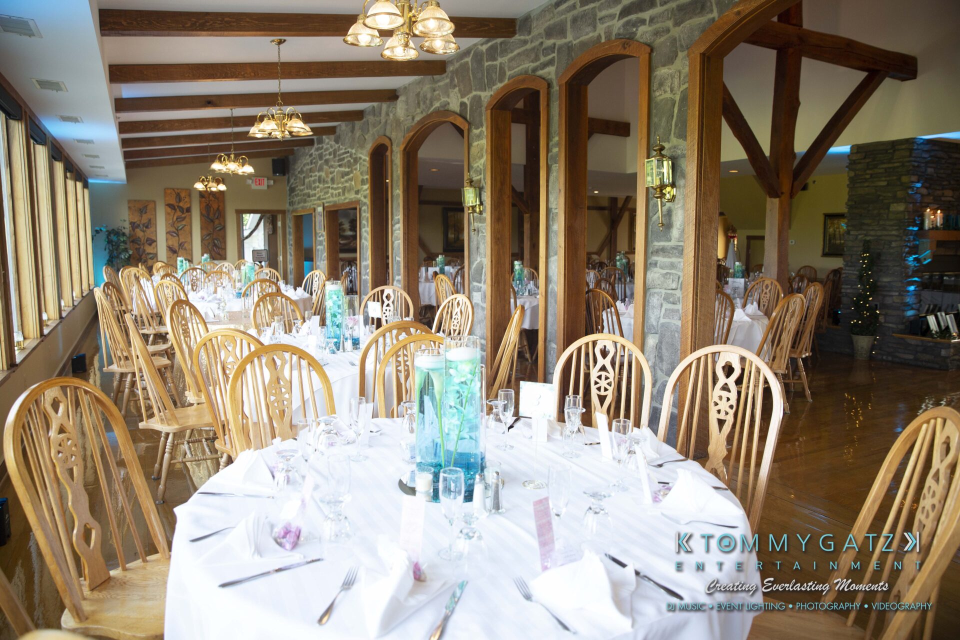 Kari-Jose-062318-RECEPTION-Low-Res-WM-419 - TOMMY GATZ ENTERTAINMENT A spacious dining hall with multiple round tables set for a banquet. Tables are covered with white tablecloths and surrounded by wooden chairs. Large windows and stone walls are visible.