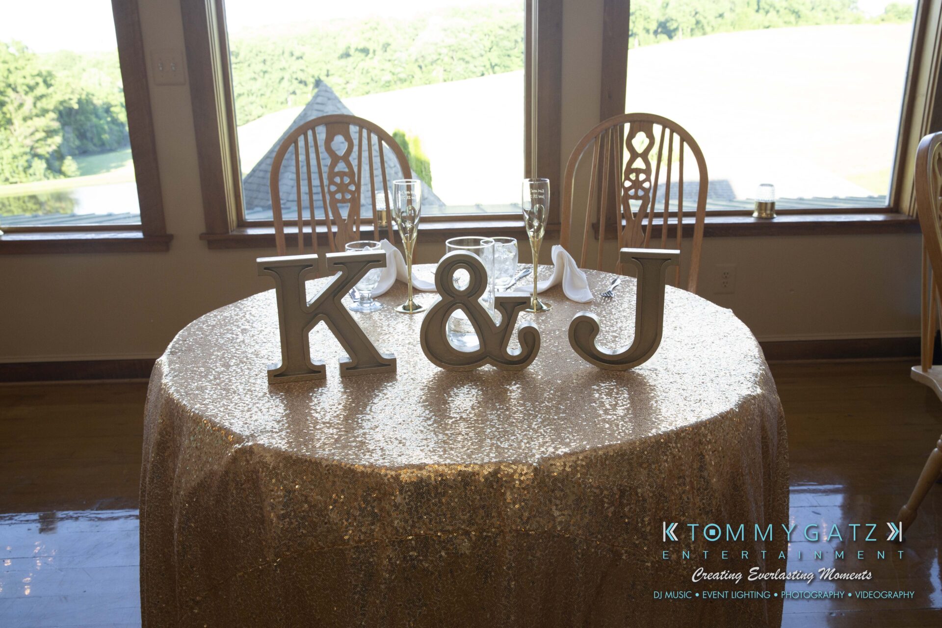 Kari-Jose-062318-RECEPTION-Low-Res-WM-423 - TOMMY GATZ ENTERTAINMENT A round, glittery table with large "K" and "J" letters, champagne glasses, napkins, and wooden chairs in a room with large windows overlooking a grassy landscape.