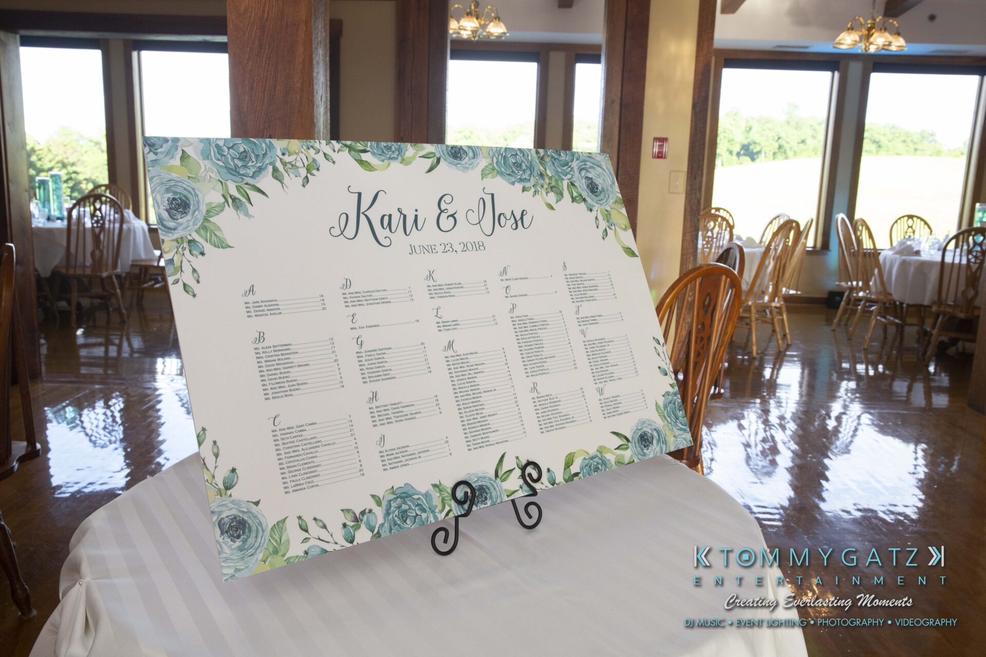 Kari-Jose-062318-RECEPTION-Low-Res-WM-424 - TOMMY GATZ ENTERTAINMENT Wedding seating chart on display with floral border and names "Kari & Joe," dated June 23, 2018, on a striped tablecloth inside a venue with wooden chairs and windows showing greenery outside.