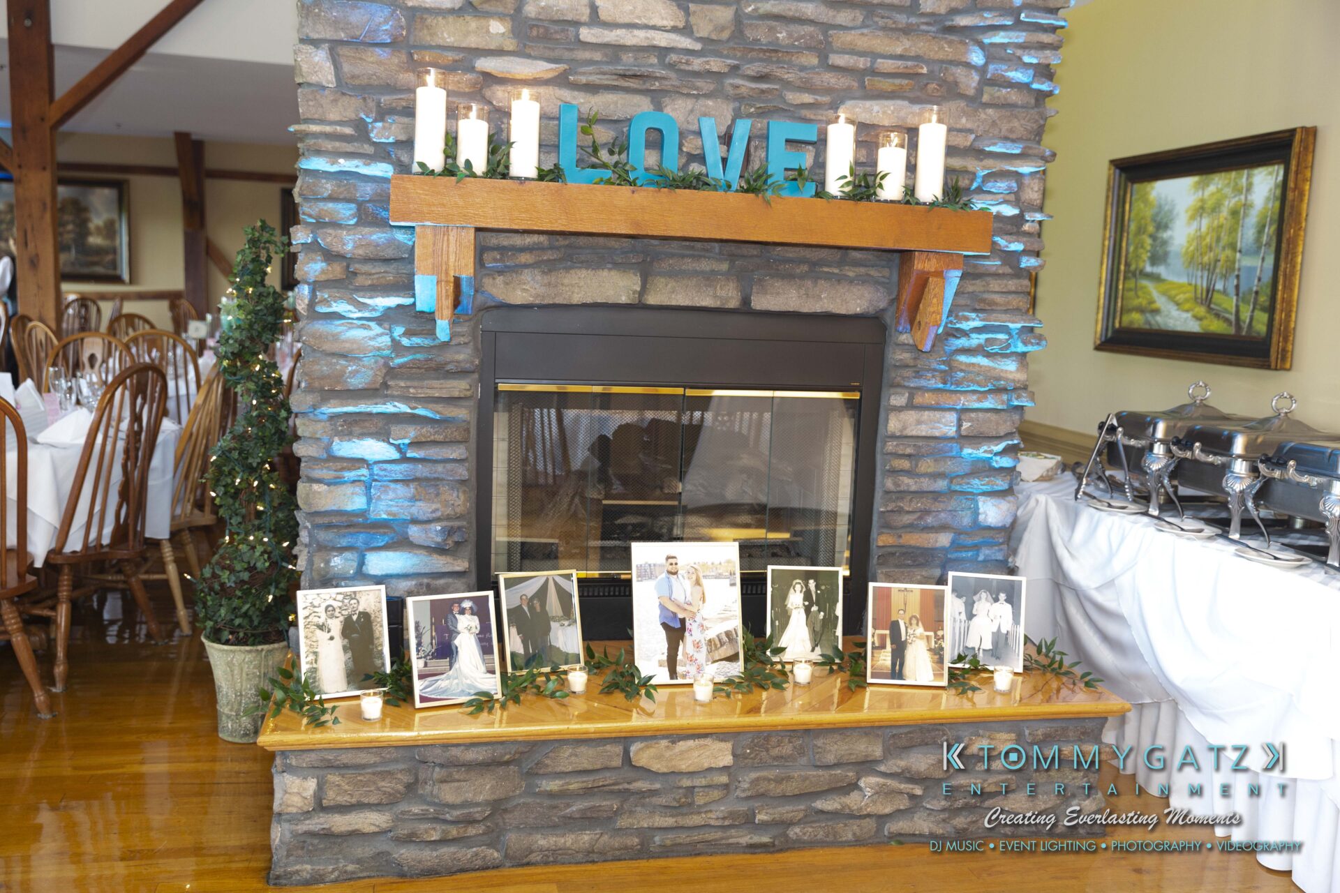 Kari-Jose-062318-RECEPTION-Low-Res-WM-426 - TOMMY GATZ ENTERTAINMENT A stone fireplace with "LOVE" in large letters. Photos of weddings are placed along the mantle, and five candles are above. Dining tables are visible in the background.