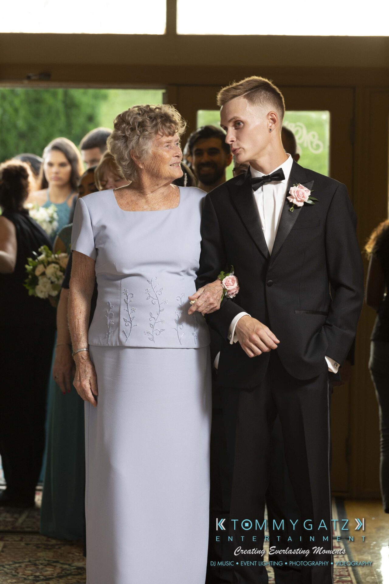 Kari-Jose-062318-RECEPTION-Low-Res-WM-436 - TOMMY GATZ ENTERTAINMENT An elderly woman and a young man in a tuxedo walking together, smiling at each other, during a formal event.