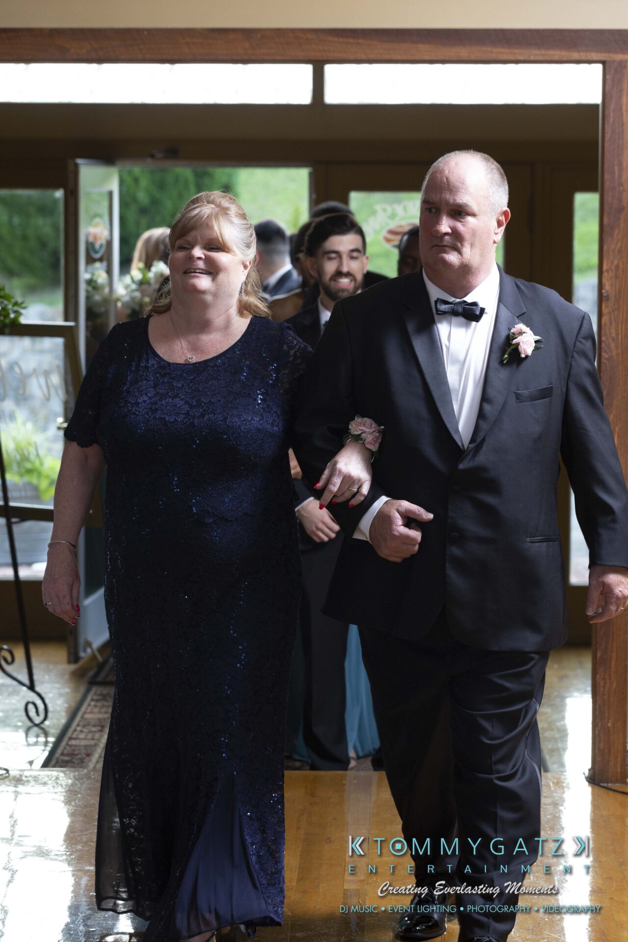 Kari-Jose-062318-RECEPTION-Low-Res-WM-439 - TOMMY GATZ ENTERTAINMENT A couple in formal attire walks arm in arm at an indoor event, with other guests visible in the background.