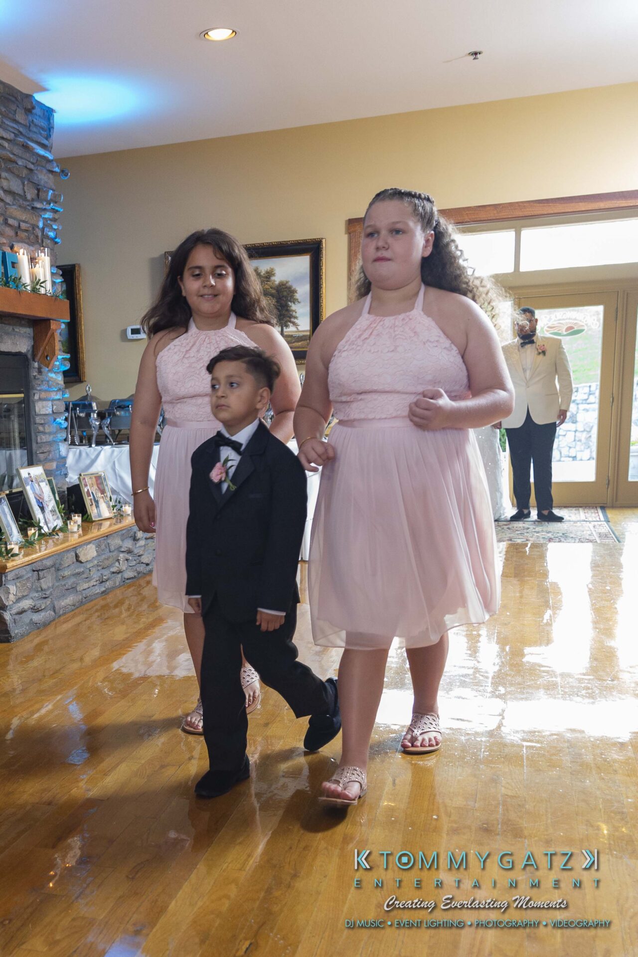 Kari-Jose-062318-RECEPTION-Low-Res-WM-449 - TOMMY GATZ ENTERTAINMENT Two young girls in pink dresses walk alongside a boy in a suit on a wooden floor indoors. A fireplace and a person in the background are visible.