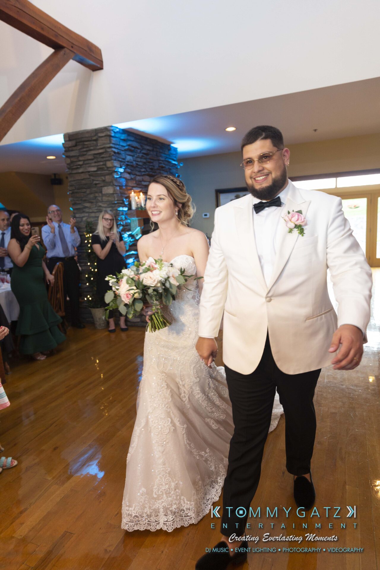 Kari-Jose-062318-RECEPTION-Low-Res-WM-453 - TOMMY GATZ ENTERTAINMENT A bride and groom walk hand-in-hand down a wooden floor aisle, smiling. The bride holds a bouquet of pink and white flowers. Onlookers are visible in the background.