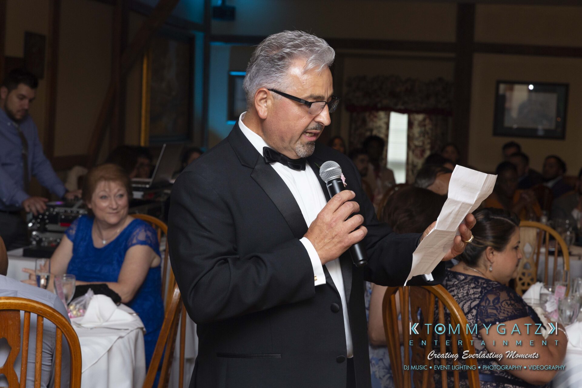 Kari-Jose-062318-RECEPTION-Low-Res-WM-456 - TOMMY GATZ ENTERTAINMENT A man in a tuxedo speaks into a microphone at a formal event, with seated guests in the background.