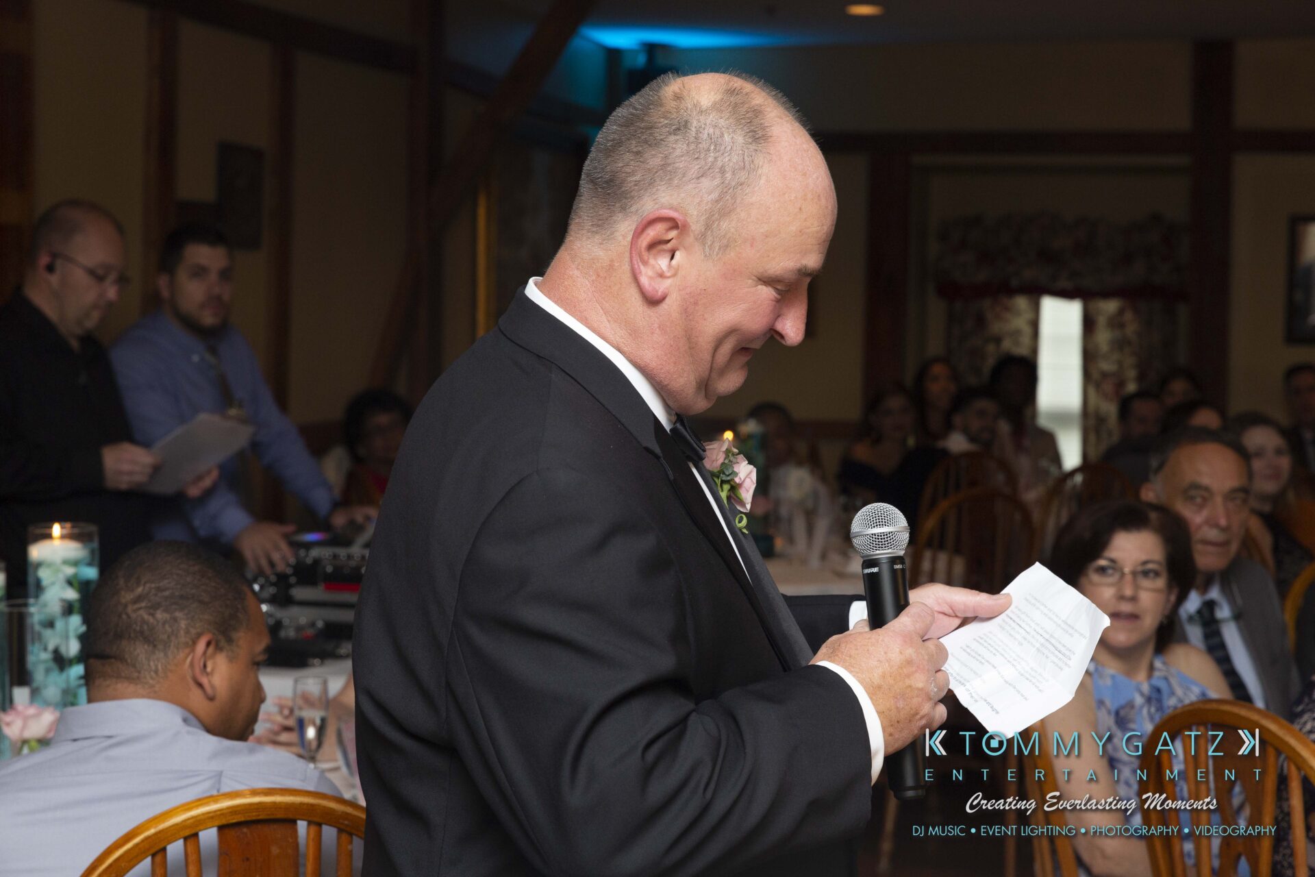 Kari-Jose-062318-RECEPTION-Low-Res-WM-463 - TOMMY GATZ ENTERTAINMENT A man in a suit speaks into a microphone while holding a piece of paper at an indoor event with seated guests in the background.