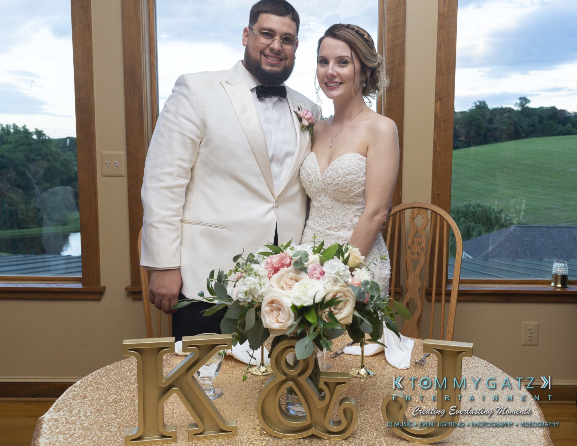 Kari-Jose-062318-RECEPTION-Low-Res-WM-508 - TOMMY GATZ ENTERTAINMENT A couple in formal attire stands behind a table with floral decorations and gold initials "K & J." A scenic landscape is visible through large windows behind them.