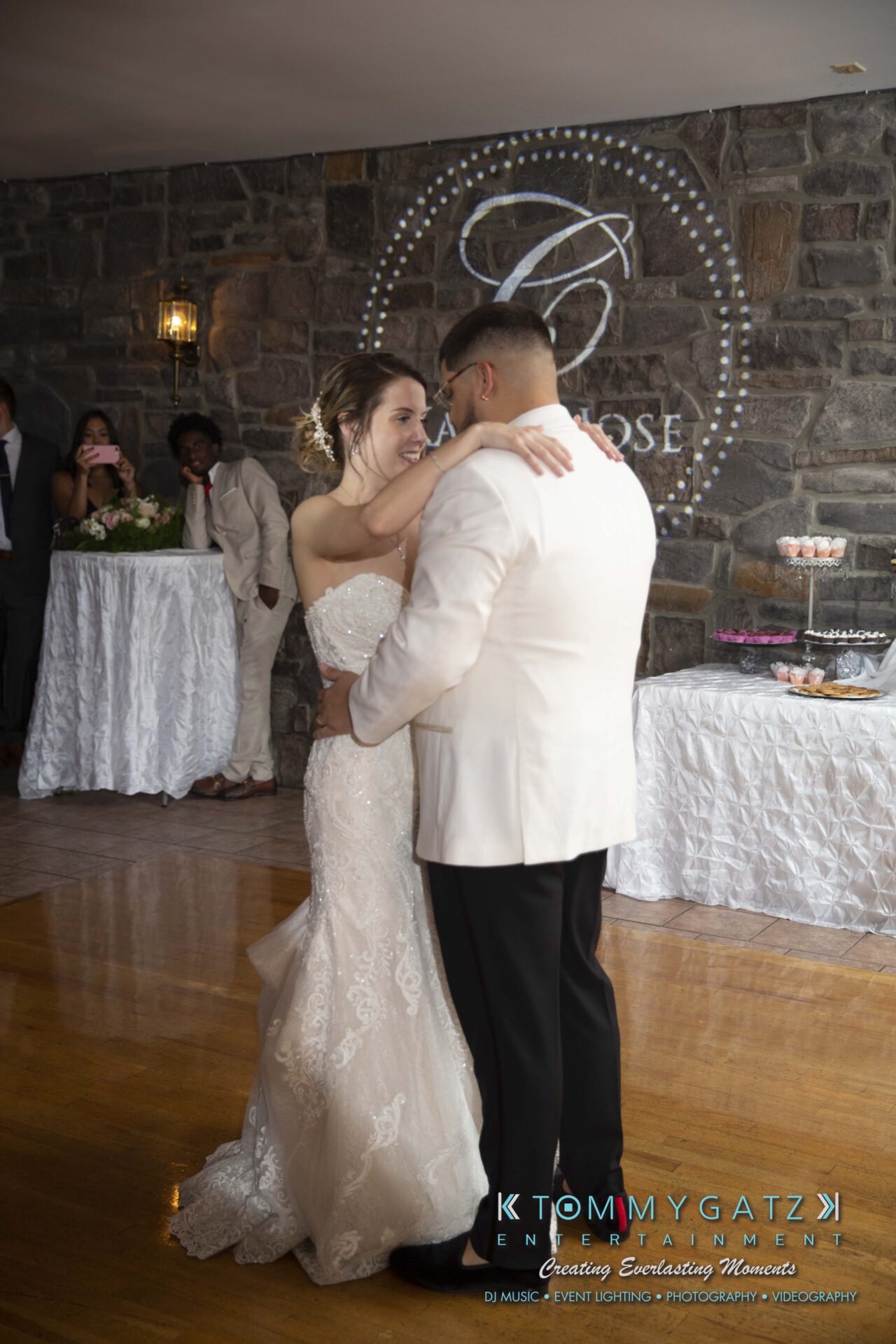 Kari-Jose-062318-RECEPTION-Low-Res-WM-515 - TOMMY GATZ ENTERTAINMENT A bride and groom share a first dance at their wedding reception, with a stone wall and a dessert table in the background.