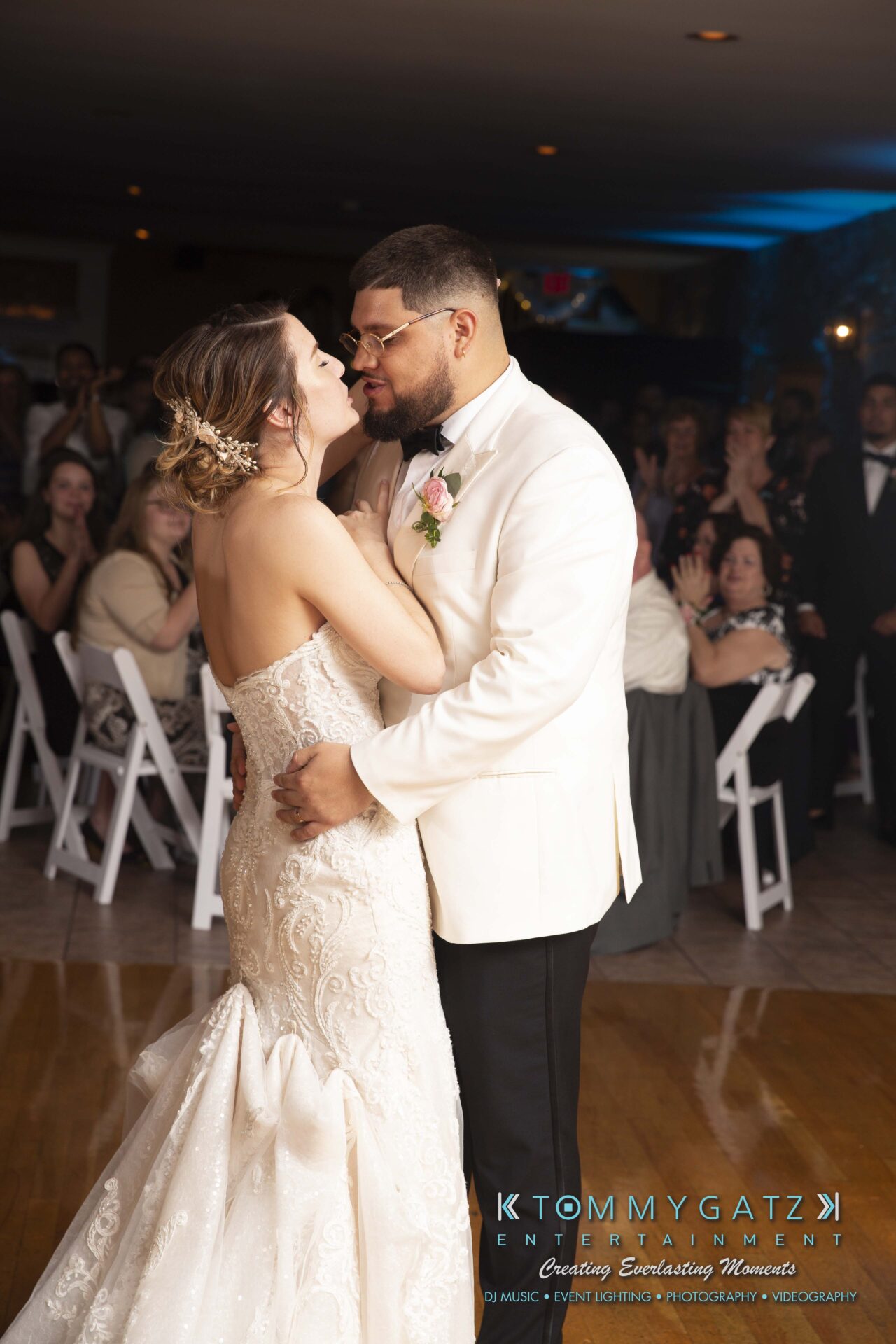 Kari-Jose-062318-RECEPTION-Low-Res-WM-523 - TOMMY GATZ ENTERTAINMENT A couple shares their first dance at a wedding reception. The bride wears a strapless lace gown, and the groom wears a white tuxedo jacket. Guests sit and stand in the background, clapping.