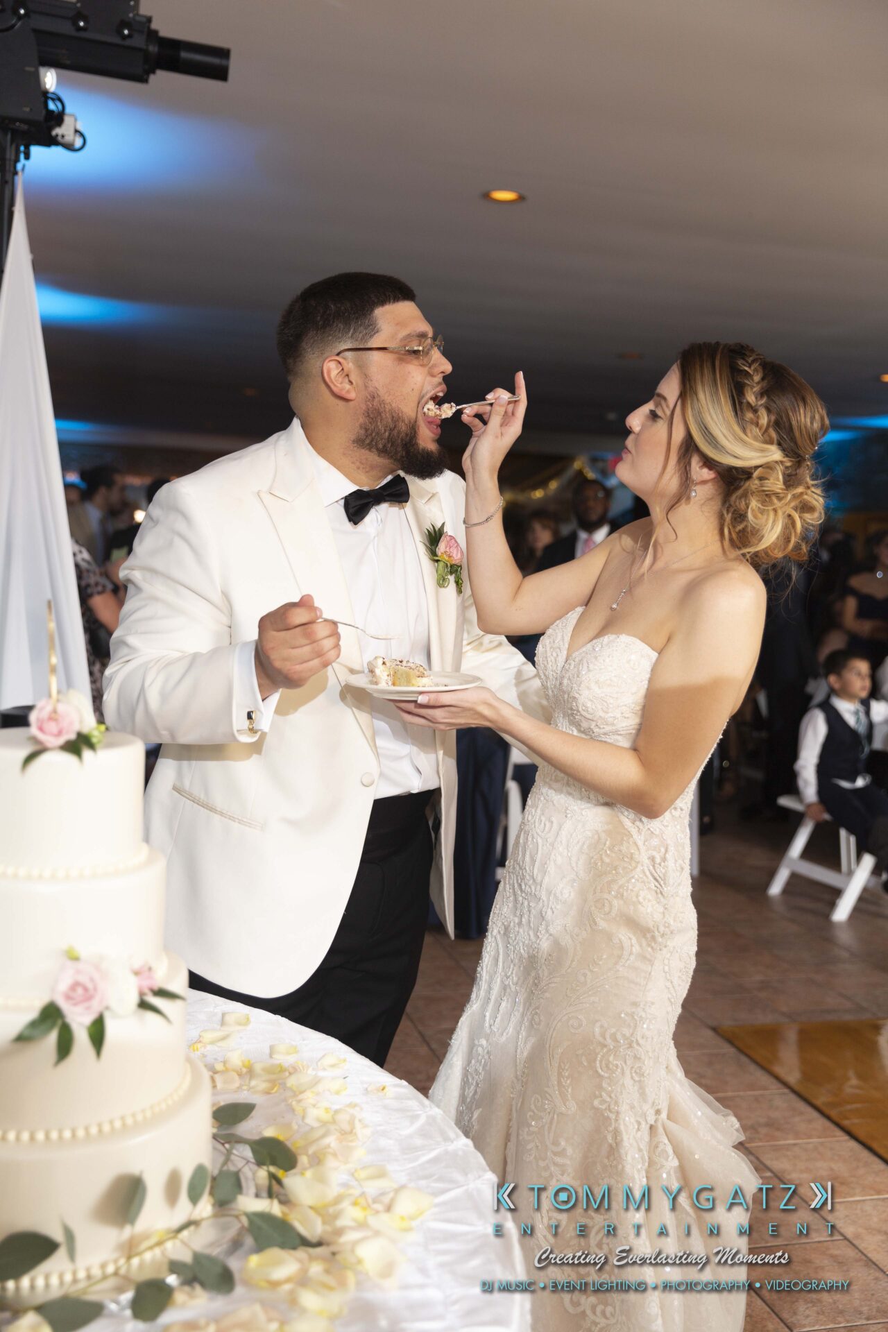 Kari-Jose-062318-RECEPTION-Low-Res-WM-555 - TOMMY GATZ ENTERTAINMENT A couple in wedding attire feeding each other cake beside a tiered cake.