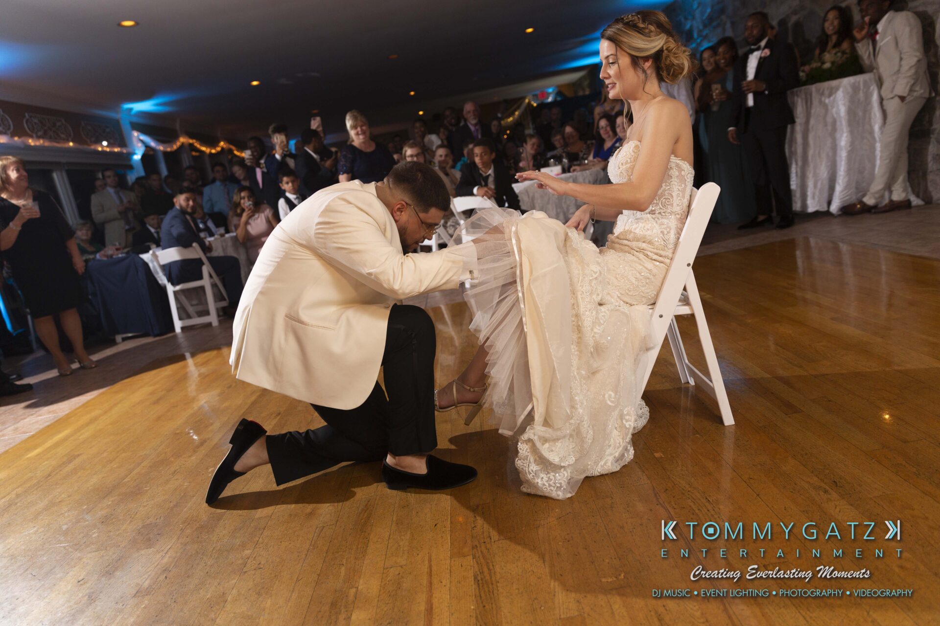 Kari-Jose-062318-RECEPTION-Low-Res-WM-573 - TOMMY GATZ ENTERTAINMENT A groom kneels in front of a bride sitting on a chair during a wedding, performing the garter removal tradition, while guests watch in the background.