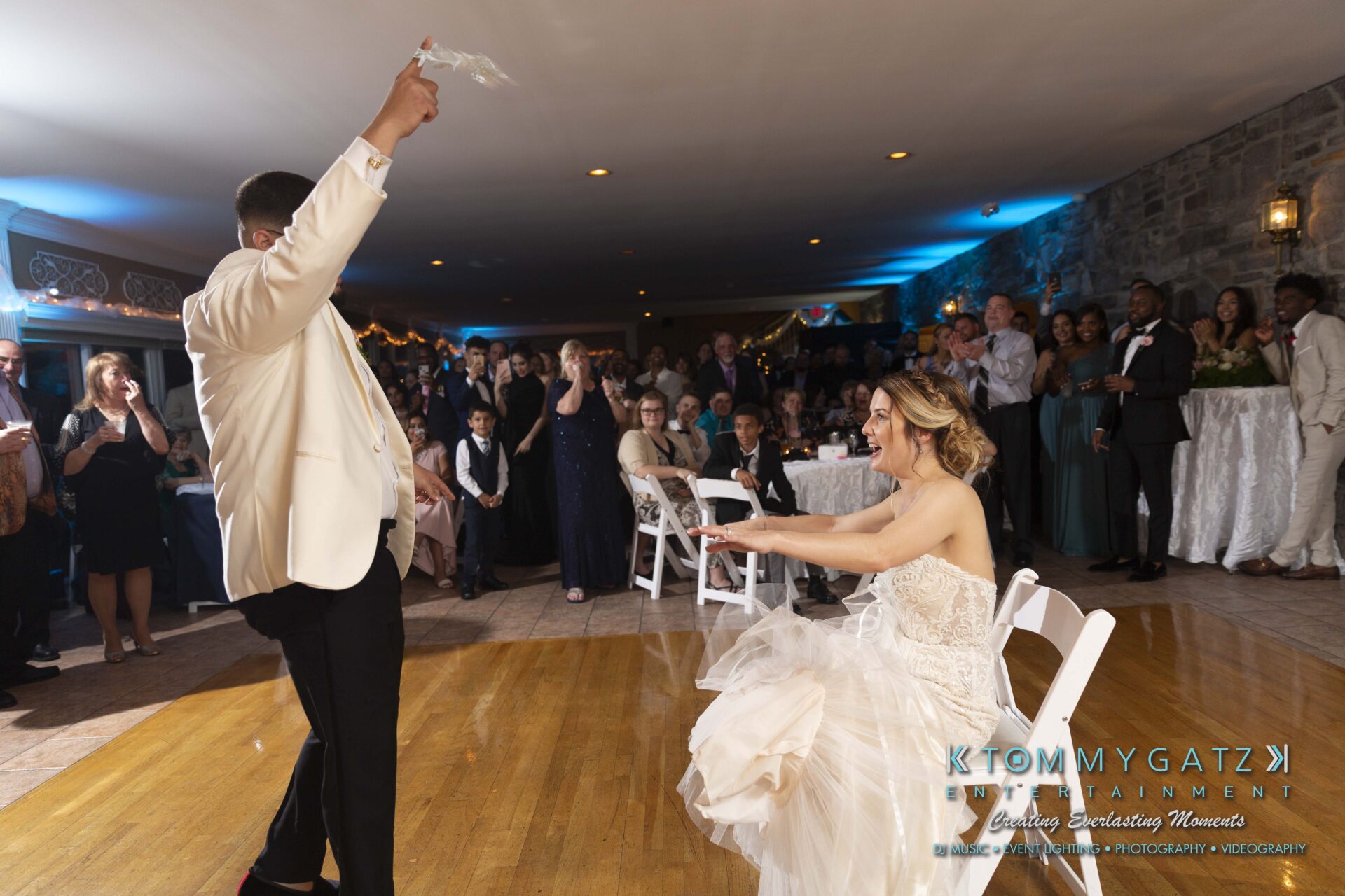 Kari-Jose-062318-RECEPTION-Low-Res-WM-580 - TOMMY GATZ ENTERTAINMENT A groom in a white jacket tosses a garter at a wedding reception. The bride sits on a chair, and guests watch from the background.