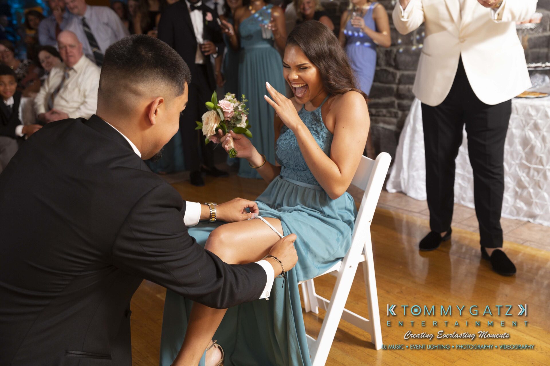 Kari-Jose-062318-RECEPTION-Low-Res-WM-595 - TOMMY GATZ ENTERTAINMENT A woman in a blue dress sits on a chair, laughing as a man kneels to remove a garter from her leg at an event. People stand and watch in the background.