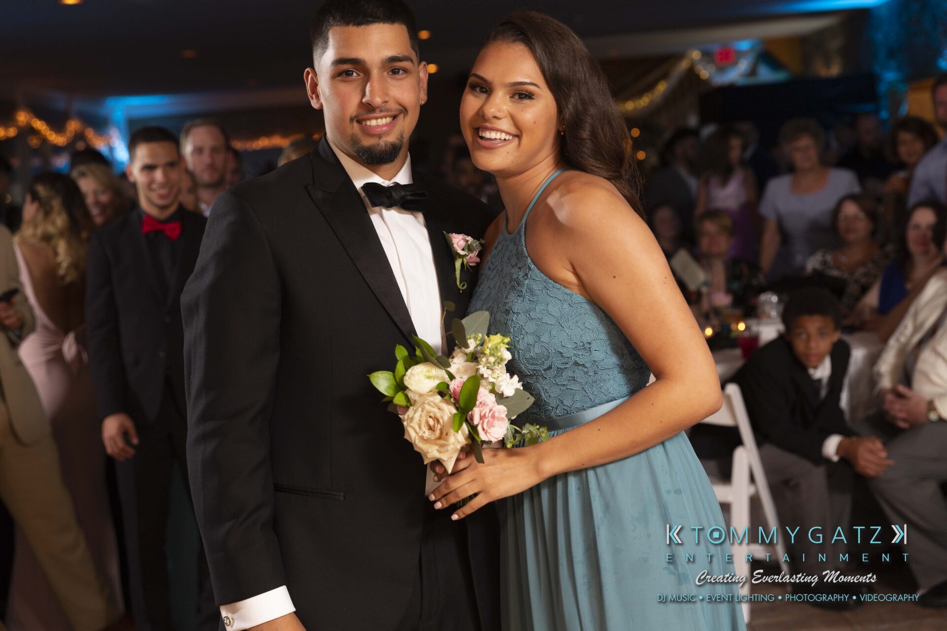Kari-Jose-062318-RECEPTION-Low-Res-WM-600 - TOMMY GATZ ENTERTAINMENT A man in a suit and a woman in a blue dress smile at the camera, holding a small bouquet, in a crowded indoor setting.