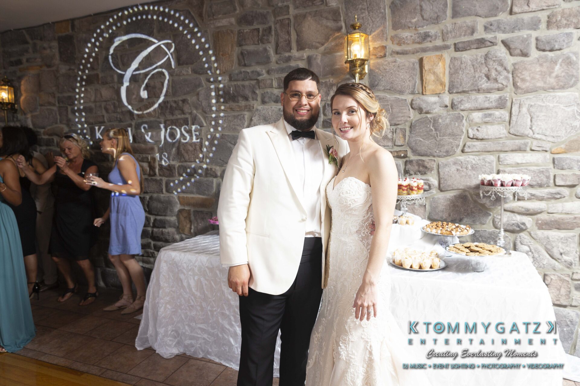 Kari-Jose-062318-RECEPTION-Low-Res-WM-601 - TOMMY GATZ ENTERTAINMENT A couple in formal attire poses in front of a dessert table at an event. A stone wall with a projected logo and a group of people in the background are also visible.