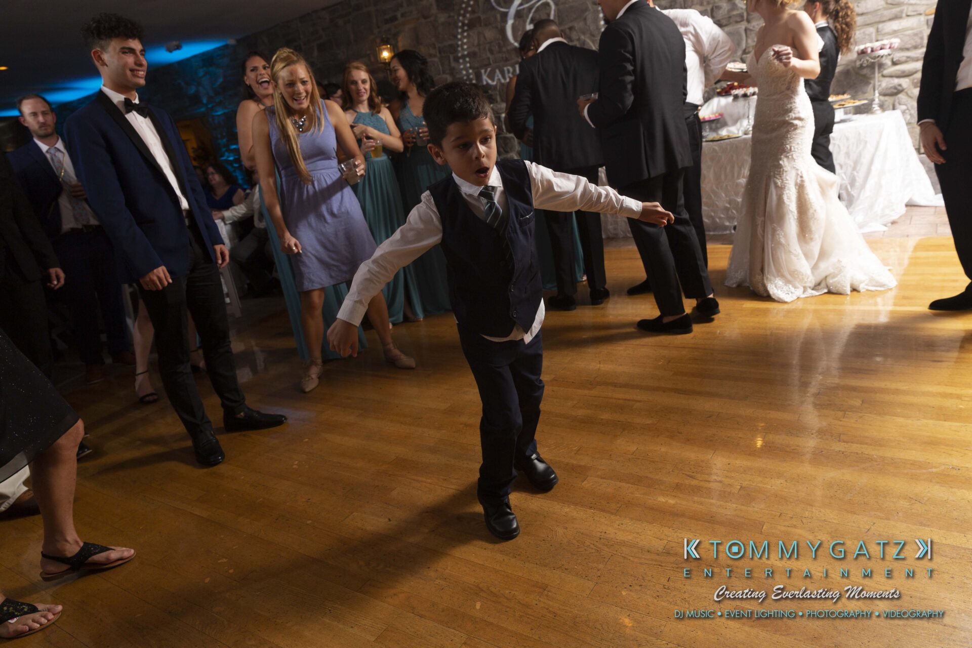 Kari-Jose-062318-RECEPTION-Low-Res-WM-604 - TOMMY GATZ ENTERTAINMENT A young boy in a suit dances energetically on a wooden floor at a formal event, surrounded by people watching and smiling.