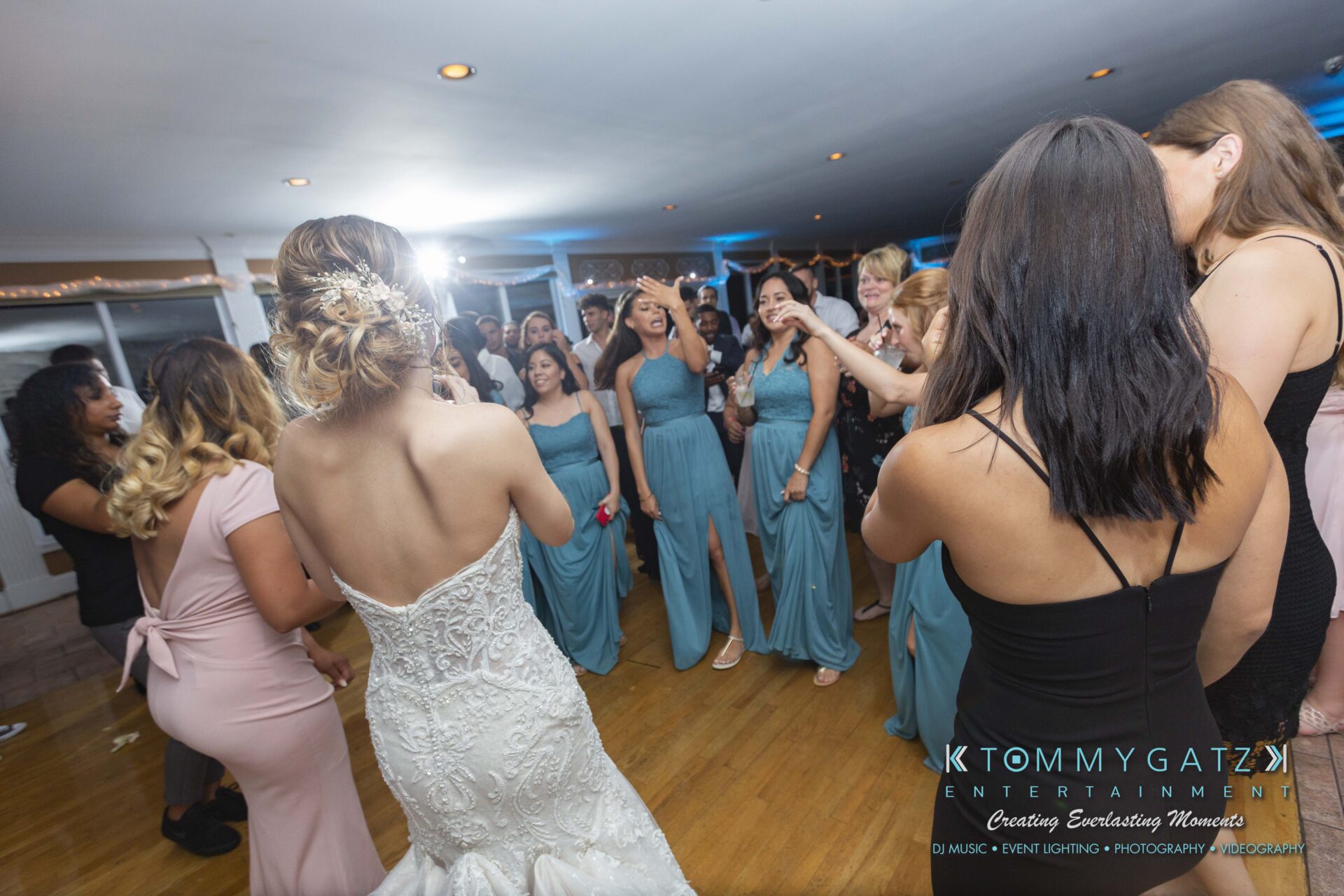 Kari-Jose-062318-RECEPTION-Low-Res-WM-659 - TOMMY GATZ ENTERTAINMENT A bride in a white gown dances with bridesmaids in teal dresses and guests in formal attire at an indoor wedding reception.