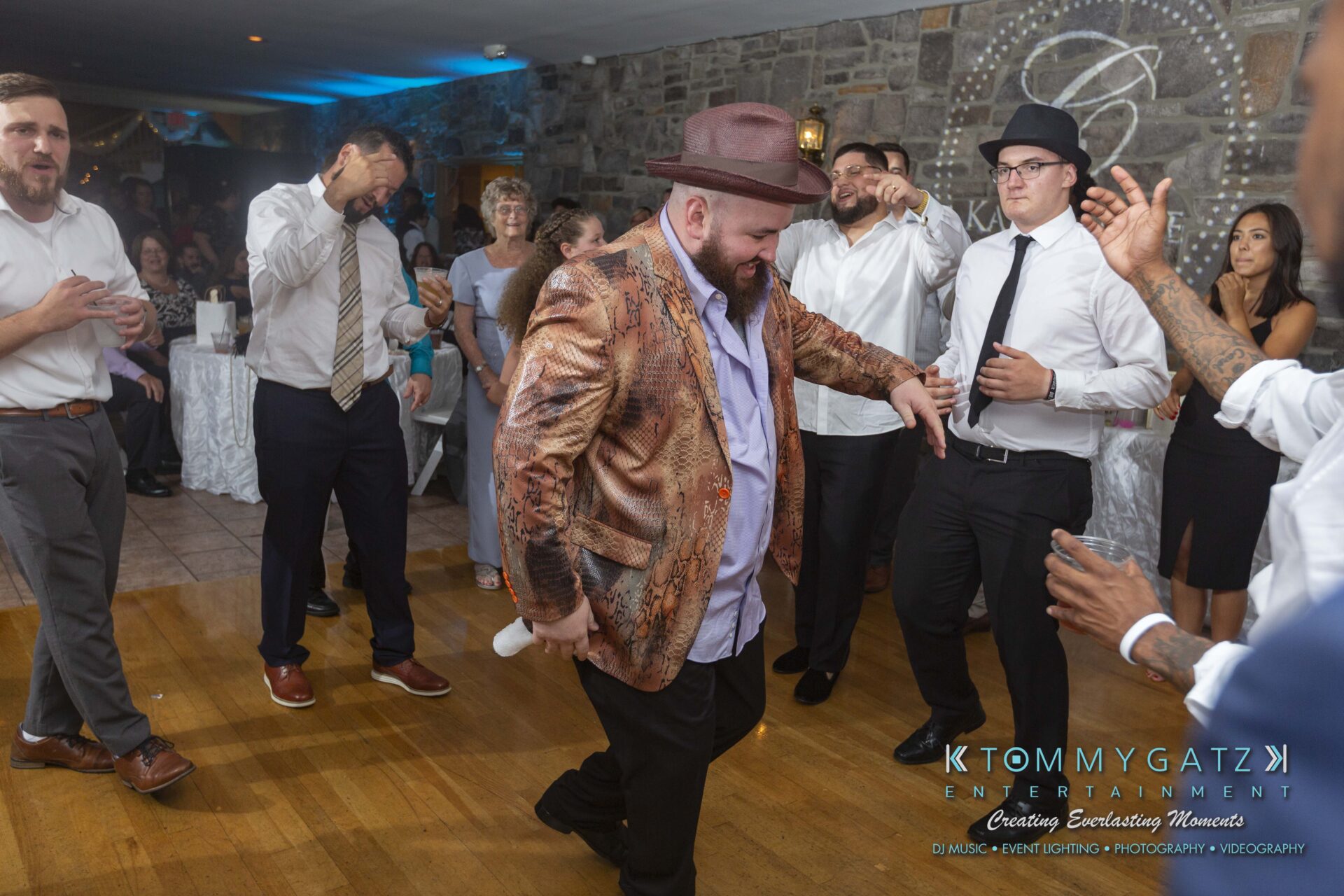 Kari-Jose-062318-RECEPTION-Low-Res-WM-675 - TOMMY GATZ ENTERTAINMENT Man in a patterned jacket and hat dances energetically on a wooden floor at an event, surrounded by casually dressed people.