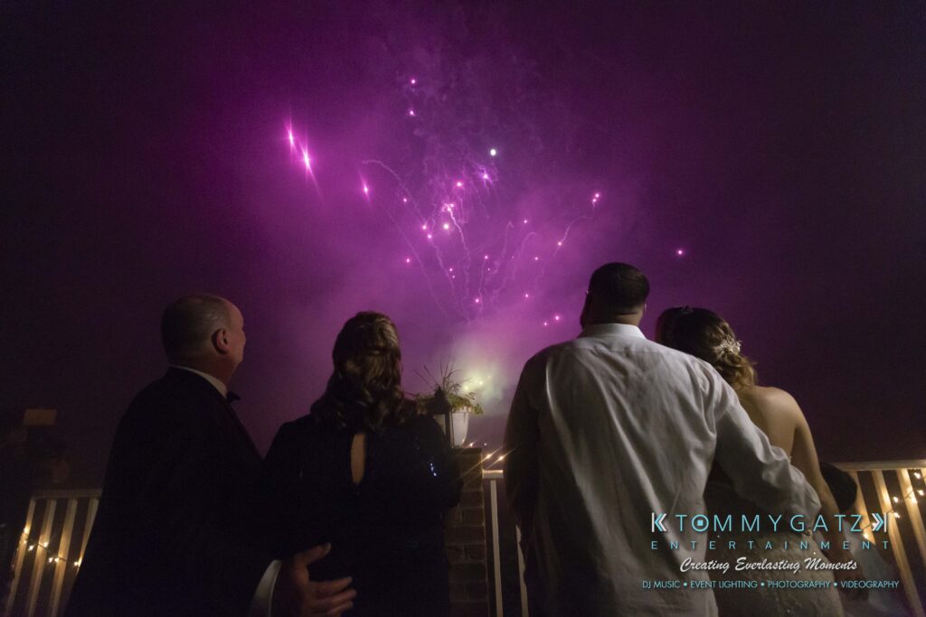 Kari-Jose-062318-RECEPTION-Low-Res-WM-704 - TOMMY GATZ ENTERTAINMENT Four people in formal attire watch pink fireworks in the night sky from a balcony.