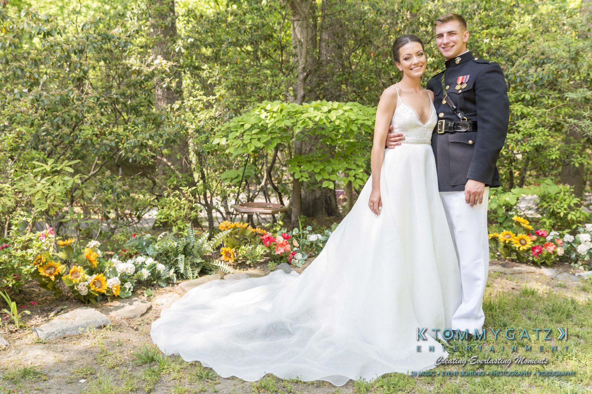 Keara-Patrick-Wedding-51218-FINALS-Low-Res-WM-1 - TOMMY GATZ ENTERTAINMENT Bride in a white gown and groom in a military uniform stand smiling in a garden, surrounded by greenery and flowers.