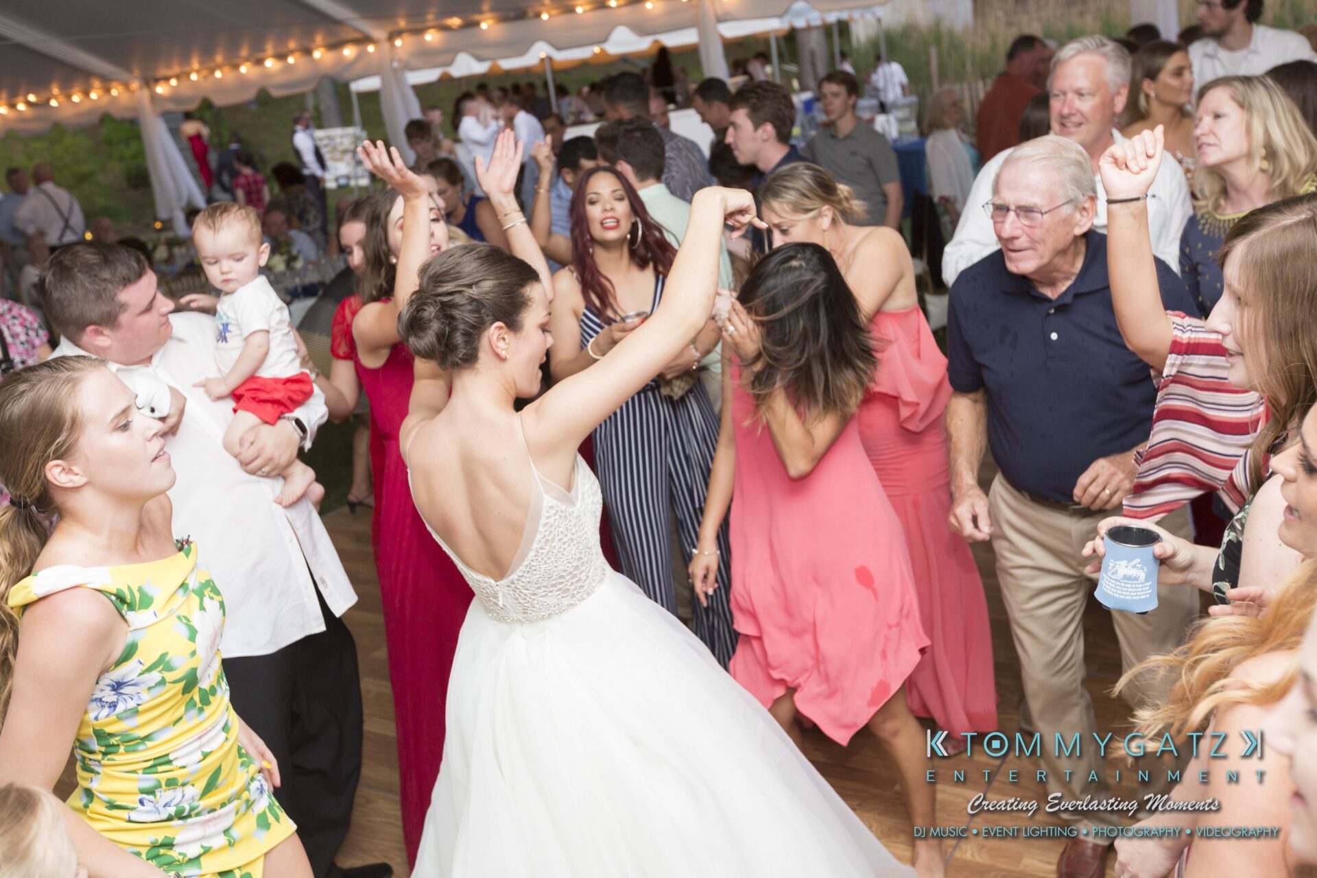 Keara-Patrick-Wedding-51218-FINALS-Low-Res-WM-13 - TOMMY GATZ ENTERTAINMENT A bride dances in a white dress among guests at an outdoor event. People are gathered around, some holding drinks, and a child is visible being held. String lights hang above.