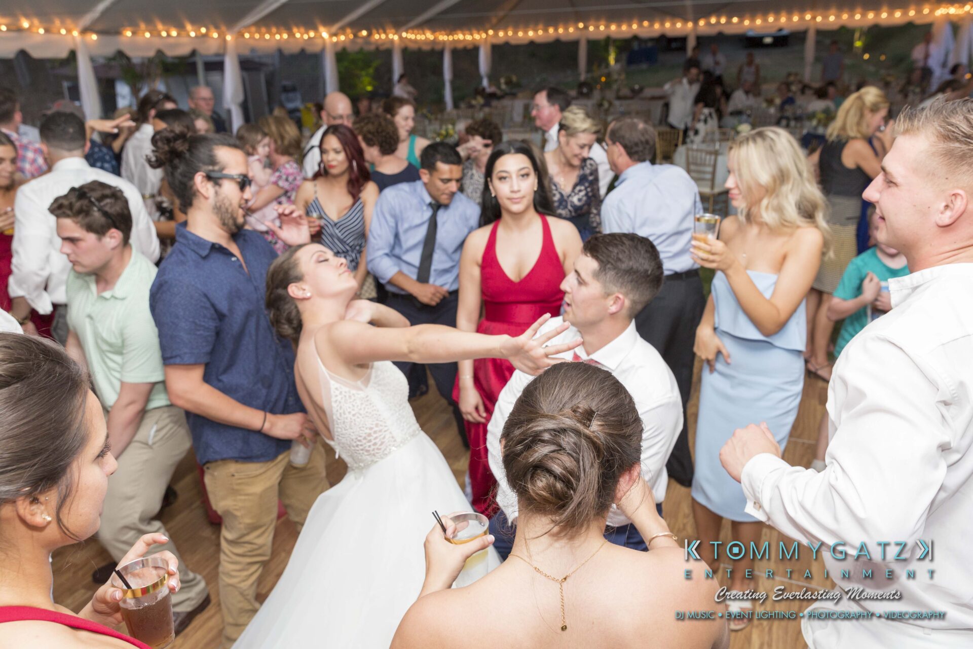 Keara-Patrick-Wedding-51218-FINALS-Low-Res-WM-14 - TOMMY GATZ ENTERTAINMENT People dancing at a wedding reception. A bride and groom are in the center, surrounded by guests under string lights.