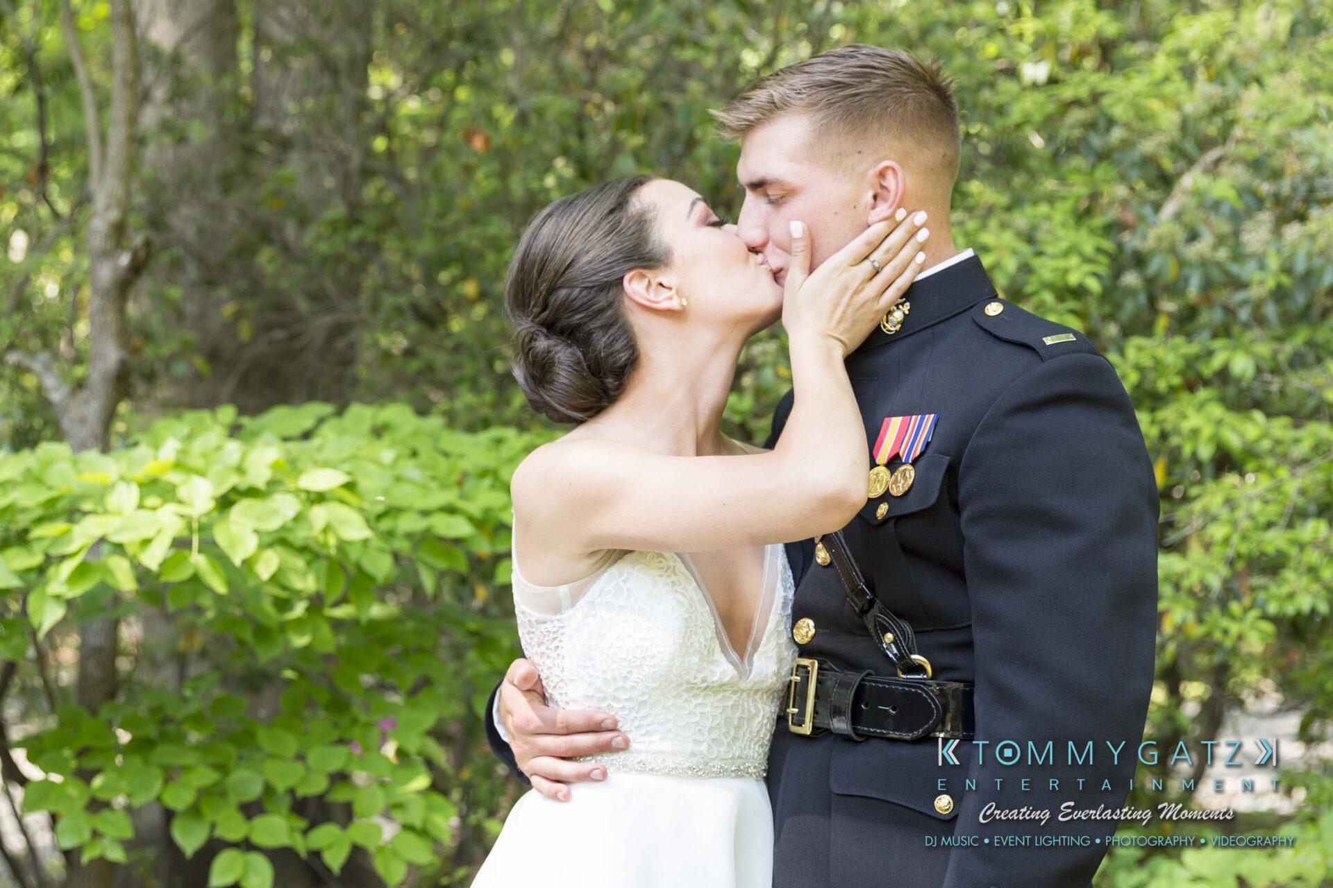 Keara-Patrick-Wedding-51218-FINALS-Low-Res-WM-2 - TOMMY GATZ ENTERTAINMENT A bride and groom in formal attire share a kiss outdoors in front of greenery. The groom wears a military dress uniform with medals.