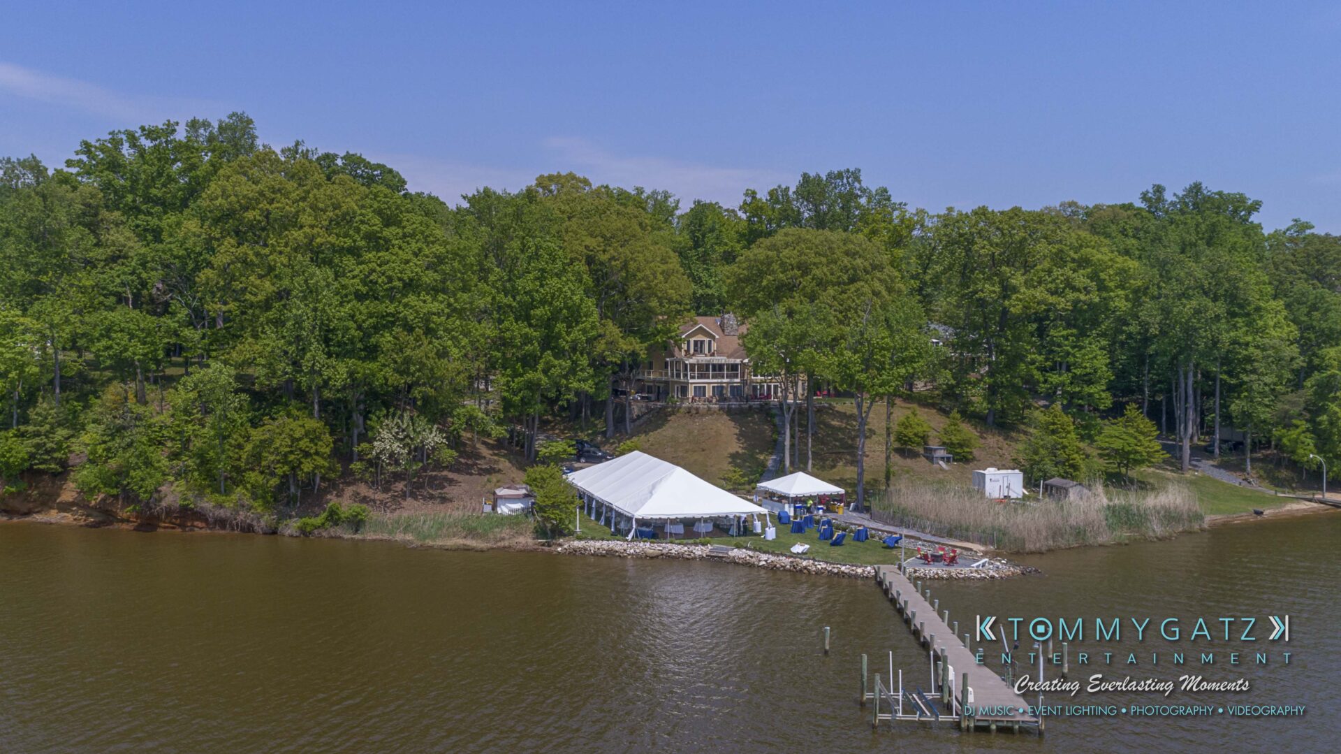 Keara-Patrick-Wedding-51218-FINALS-Low-Res-WM-21 - TOMMY GATZ ENTERTAINMENT A house with large windows is surrounded by trees and overlooks a lake. Two white tents are set up near a dock extending into the water.