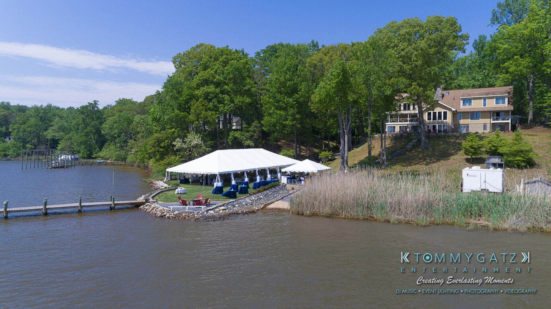 Keara-Patrick-Wedding-51218-FINALS-Low-Res-WM-22 - TOMMY GATZ ENTERTAINMENT A large white tent set up by a lake with trees and two houses in the background. A dock extends from the shoreline.