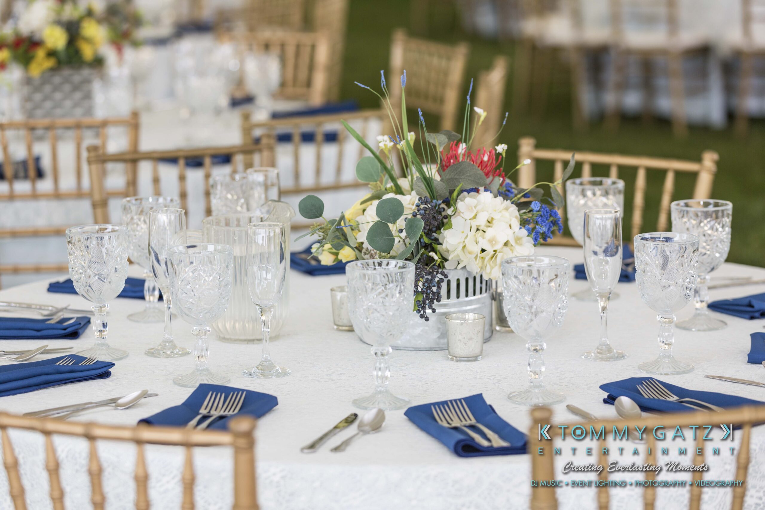 Keara-Patrick-Wedding-51218-FINALS-Low-Res-WM-3 - TOMMY GATZ ENTERTAINMENT Elegant table setting with crystal glassware, silver cutlery, blue napkins, and a floral centerpiece on a white tablecloth. Brown chairs surround the table.