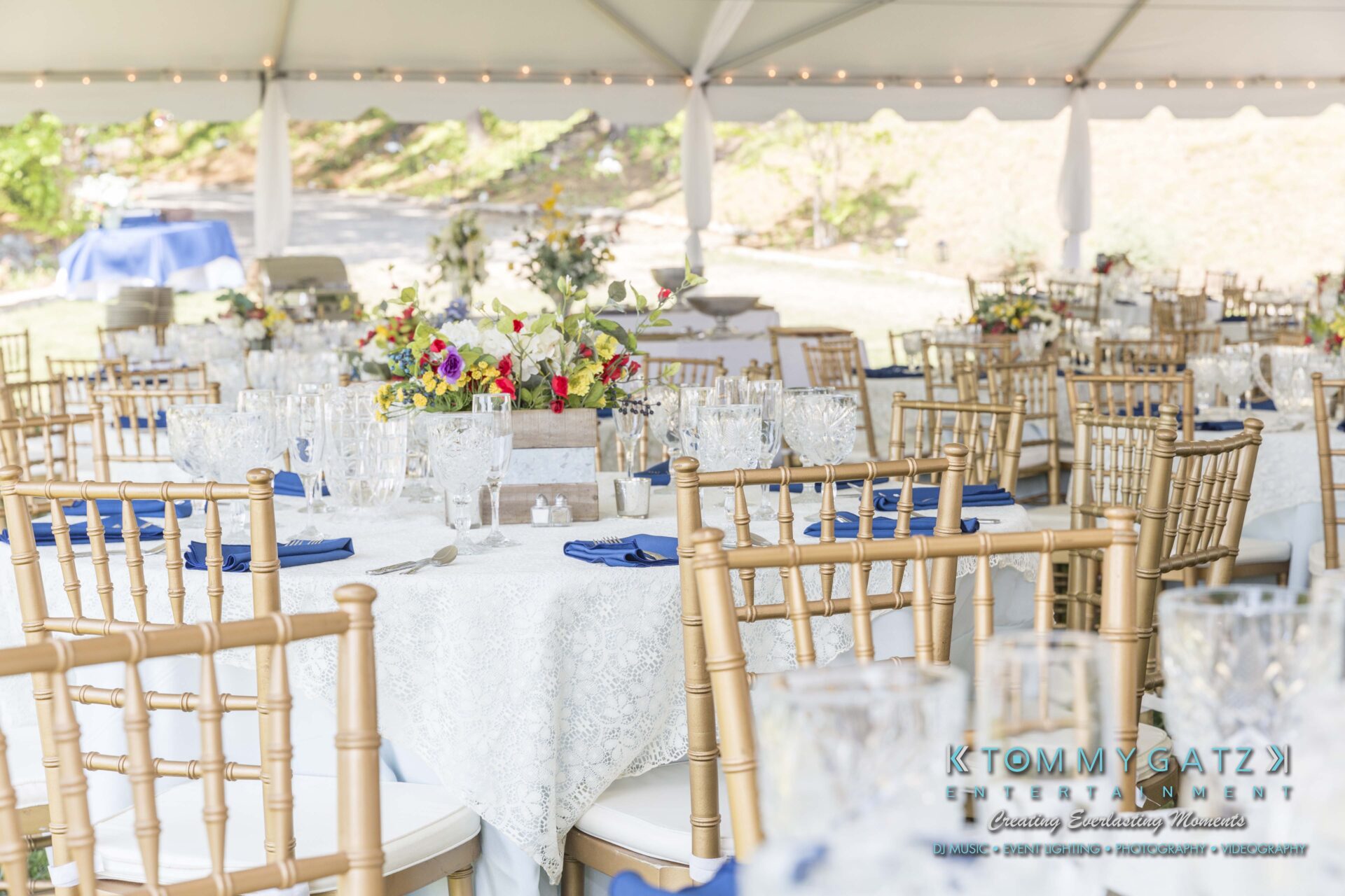 Keara-Patrick-Wedding-51218-FINALS-Low-Res-WM-6 - TOMMY GATZ ENTERTAINMENT Outdoor wedding reception setup under a canopy with round tables, white tablecloths, blue napkins, gold chairs, and floral centerpieces.