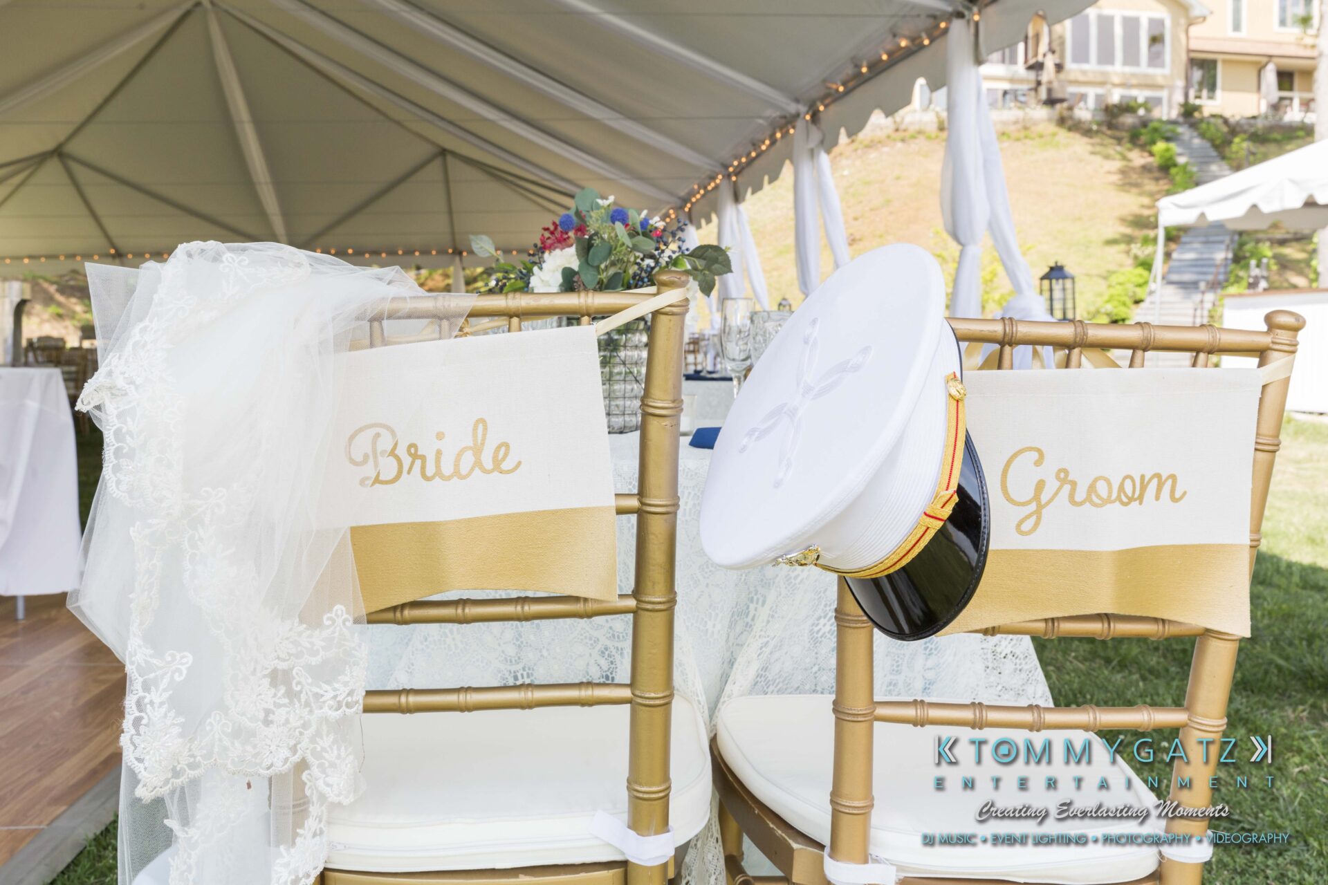 Keara-Patrick-Wedding-51218-FINALS-Low-Res-WM-7 - TOMMY GATZ ENTERTAINMENT Wedding chairs labeled "Bride" and "Groom" under a tent, with a bridal veil and military hat placed on them.