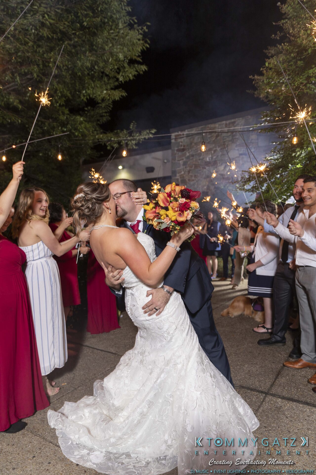 Lisa-Robert-092218-BLOG-selections-WM-102 - TOMMY GATZ ENTERTAINMENT A bride and groom kiss surrounded by guests holding sparklers. The bride holds a bouquet of red and yellow flowers.