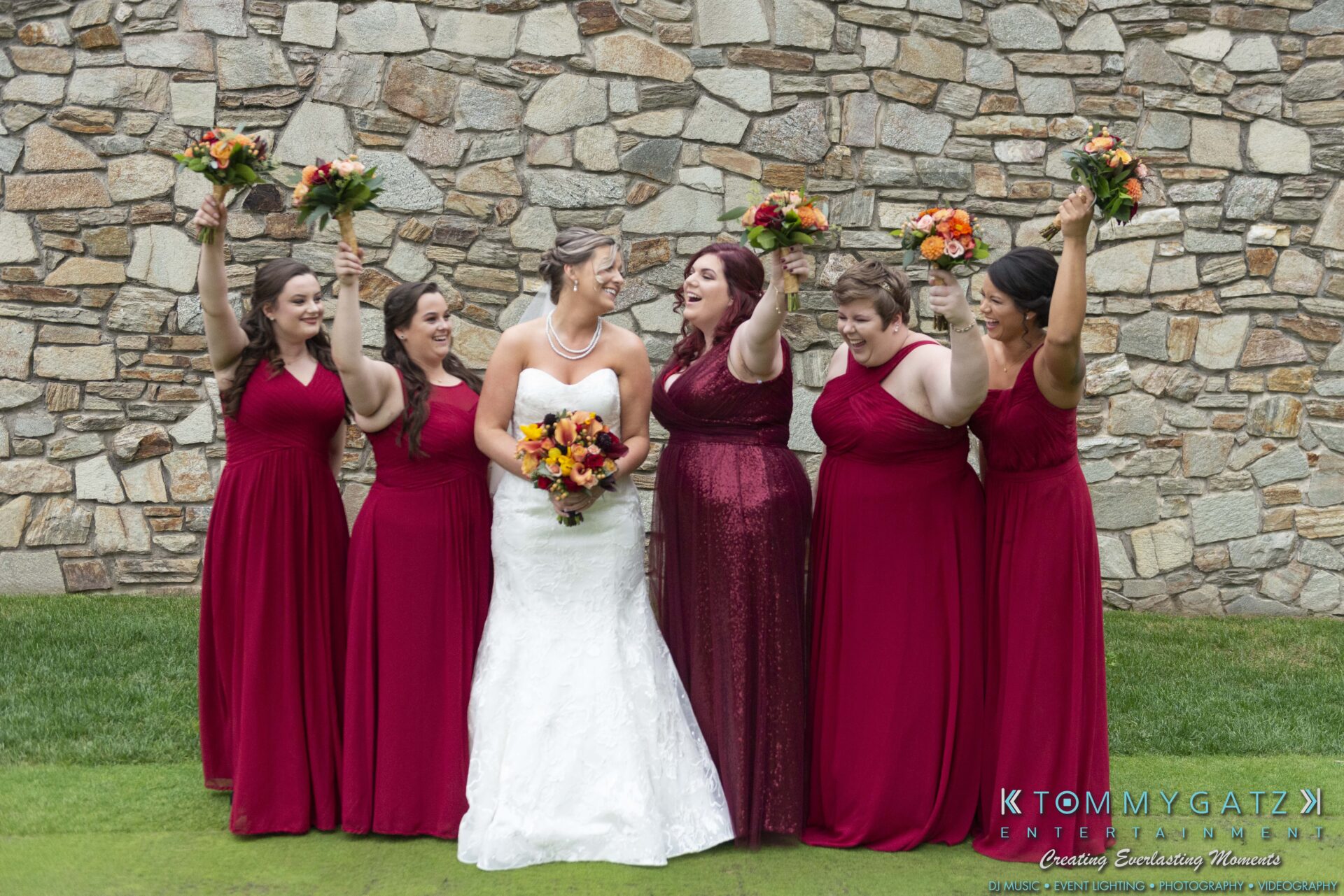 Lisa-Robert-092218-BLOG-selections-WM-11 - TOMMY GATZ ENTERTAINMENT A bride in a white dress stands with five bridesmaids in red dresses holding bouquets, smiling in front of a stone wall.