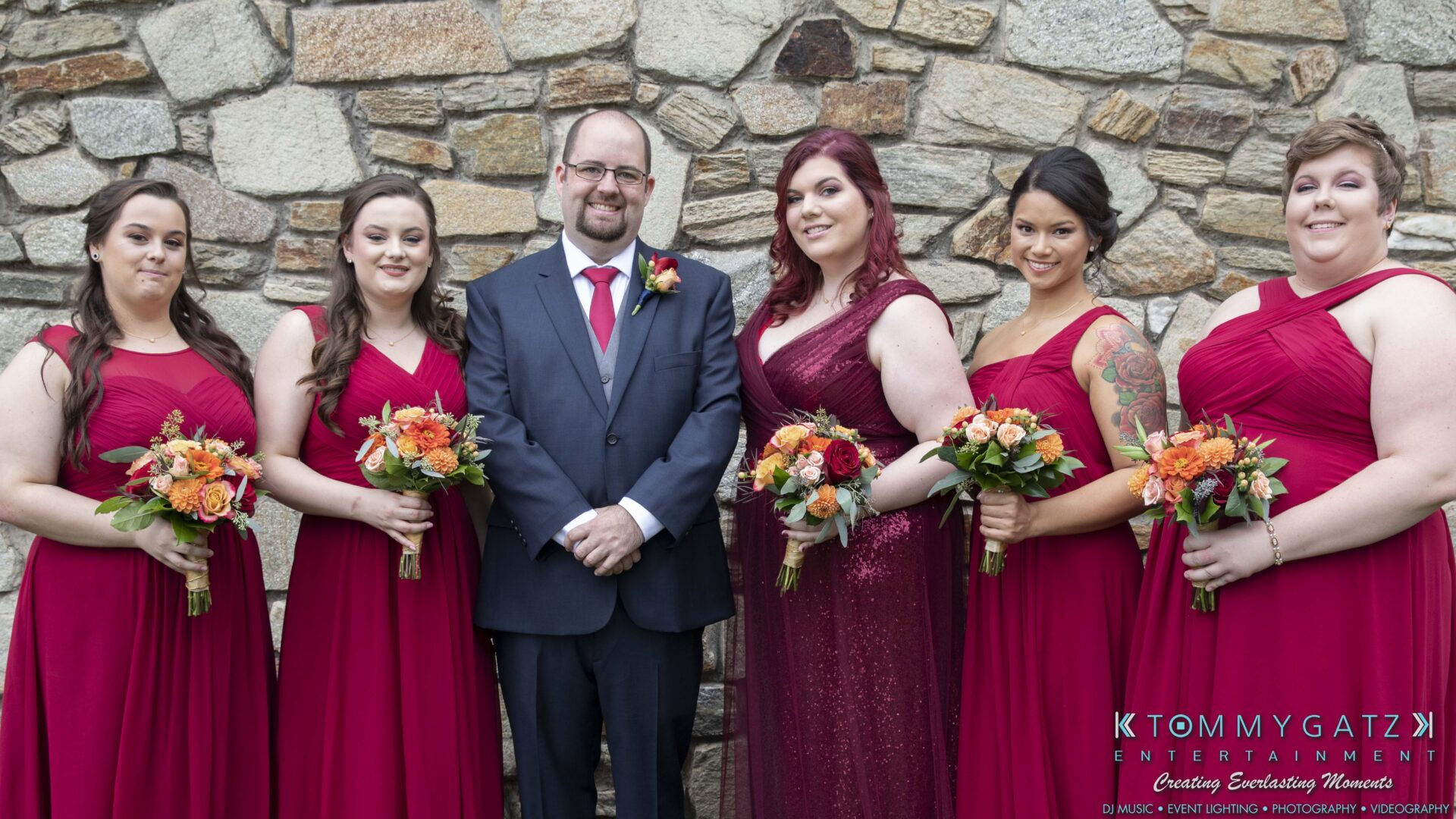 Lisa-Robert-092218-BLOG-selections-WM-15 - TOMMY GATZ ENTERTAINMENT A group photo of a groom in a suit and five bridesmaids in red dresses holding bouquets, standing against a stone wall.