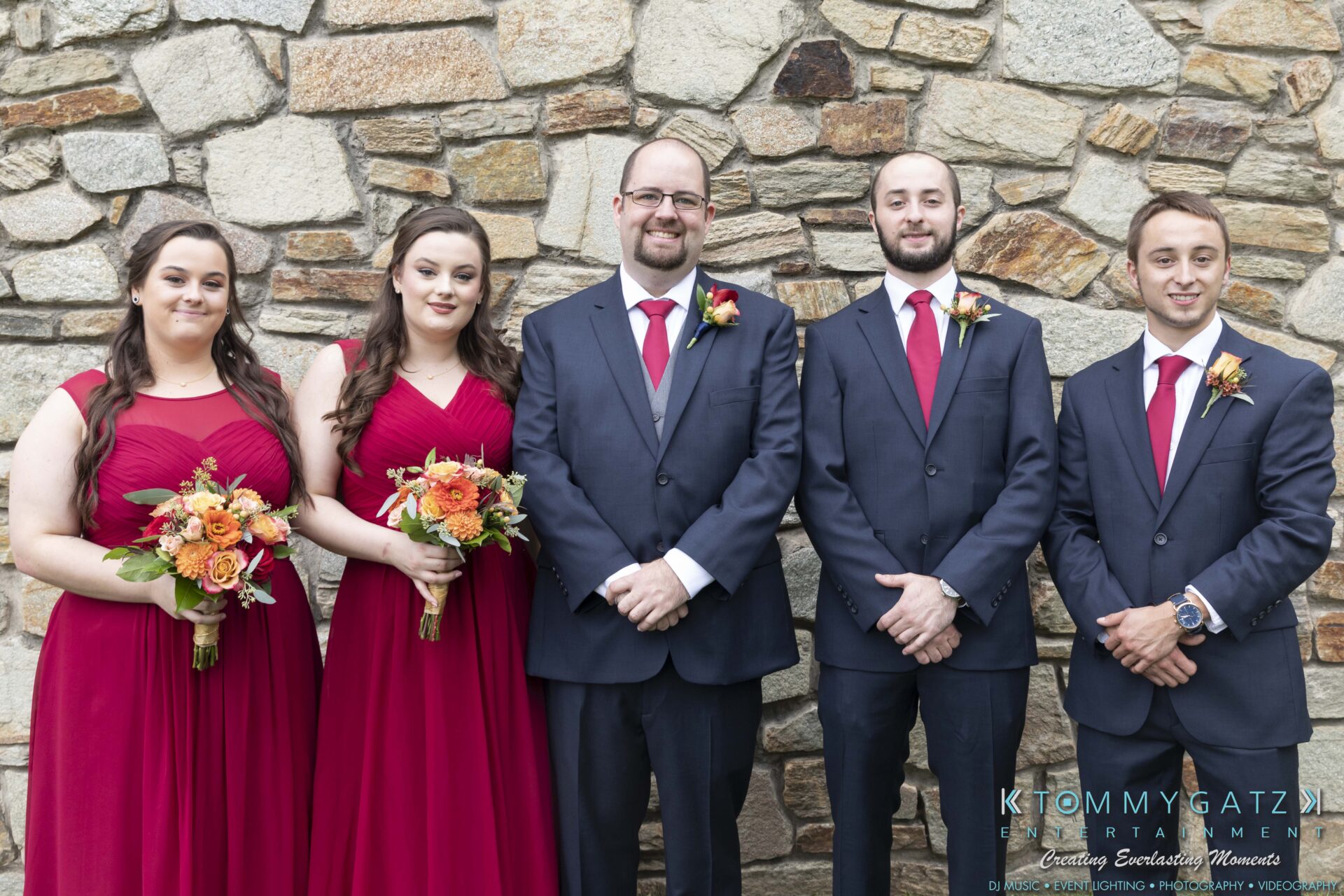 Lisa-Robert-092218-BLOG-selections-WM-16 - TOMMY GATZ ENTERTAINMENT Five people in formal attire stand in front of a stone wall. Three women wear red dresses and hold bouquets. Two men wear dark suits with red ties.