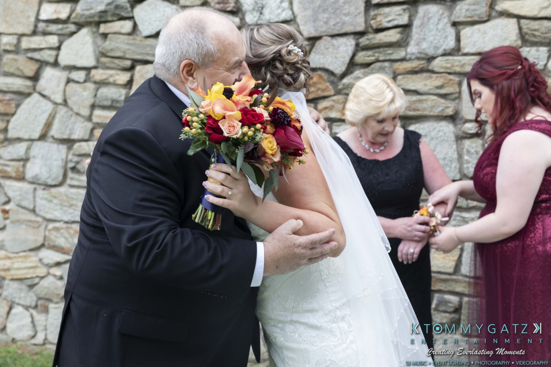 Lisa-Robert-092218-BLOG-selections-WM-22 - TOMMY GATZ ENTERTAINMENT A bride in a white gown hugs a man in a suit holding a vibrant bouquet. Two women, one in black and one in red, stand nearby. A stone wall is in the background.