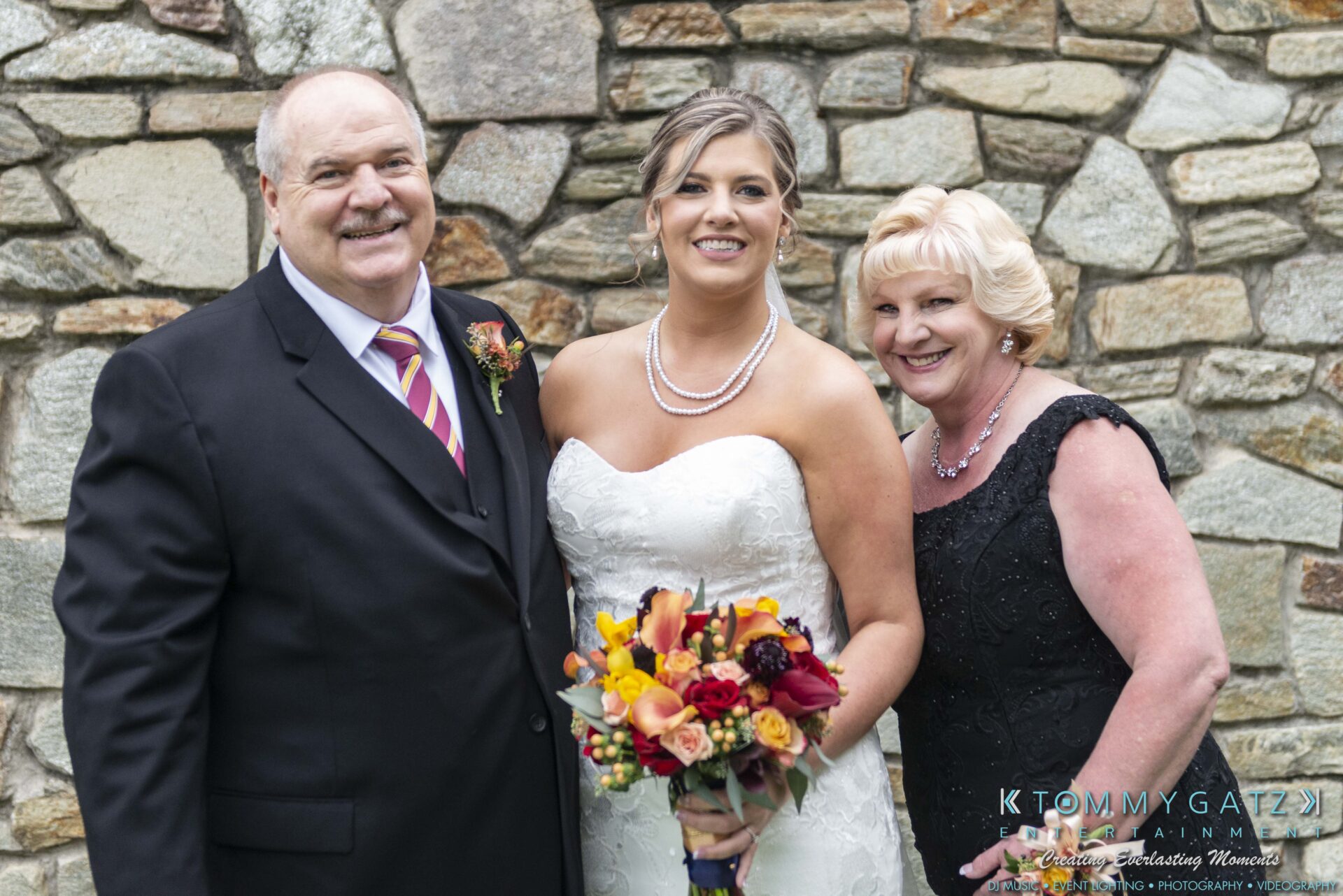 Lisa-Robert-092218-BLOG-selections-WM-23 - TOMMY GATZ ENTERTAINMENT A bride in a white gown holds a bouquet, standing between two older adults in formal attire, in front of a stone wall.