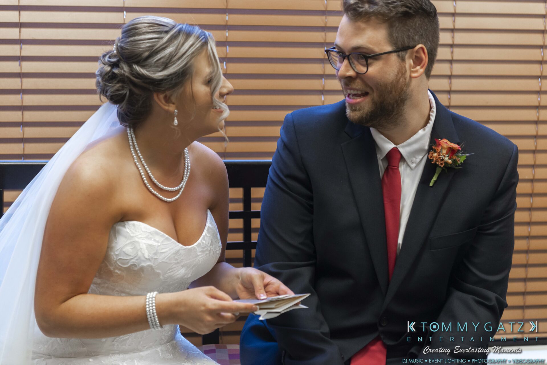 Lisa-Robert-092218-BLOG-selections-WM-27 - TOMMY GATZ ENTERTAINMENT A bride in a white dress and veil sits beside a groom in a dark suit and red tie. They are smiling and holding notes, seated in front of window blinds.