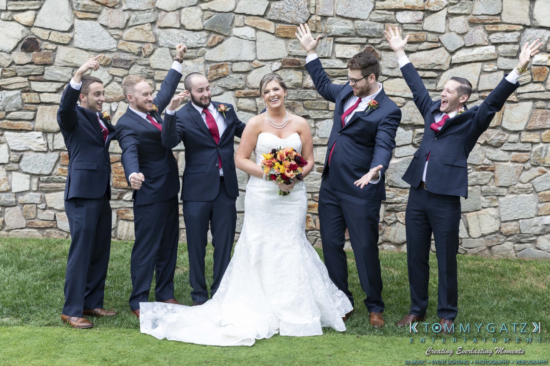 Lisa-Robert-092218-BLOG-selections-WM-30 1 - TOMMY GATZ ENTERTAINMENT A bride in a white gown holds a bouquet, surrounded by five groomsmen in dark suits with pink ties. They smile and raise their hands while standing in front of a stone wall on grass.
