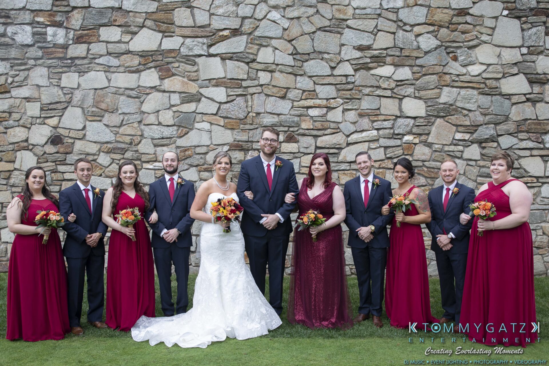 Lisa-Robert-092218-BLOG-selections-WM-32 - TOMMY GATZ ENTERTAINMENT A wedding party with the bride and groom in the center, bridesmaids in red dresses, and groomsmen in dark suits, standing in front of a stone wall.