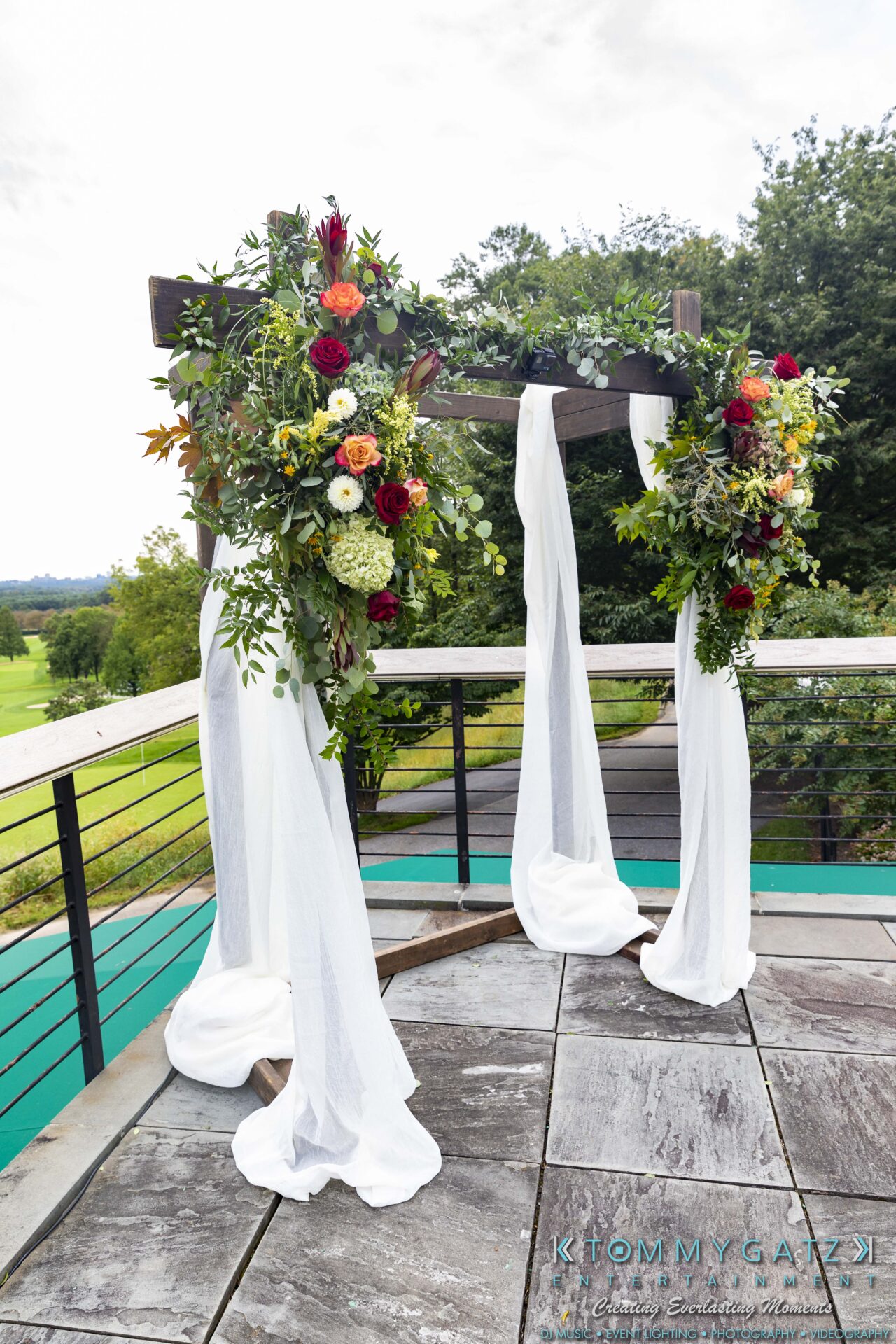 Lisa-Robert-092218-BLOG-selections-WM-37 - TOMMY GATZ ENTERTAINMENT Wedding arch decorated with colorful flowers and white fabric, set on an outdoor deck overlooking greenery.