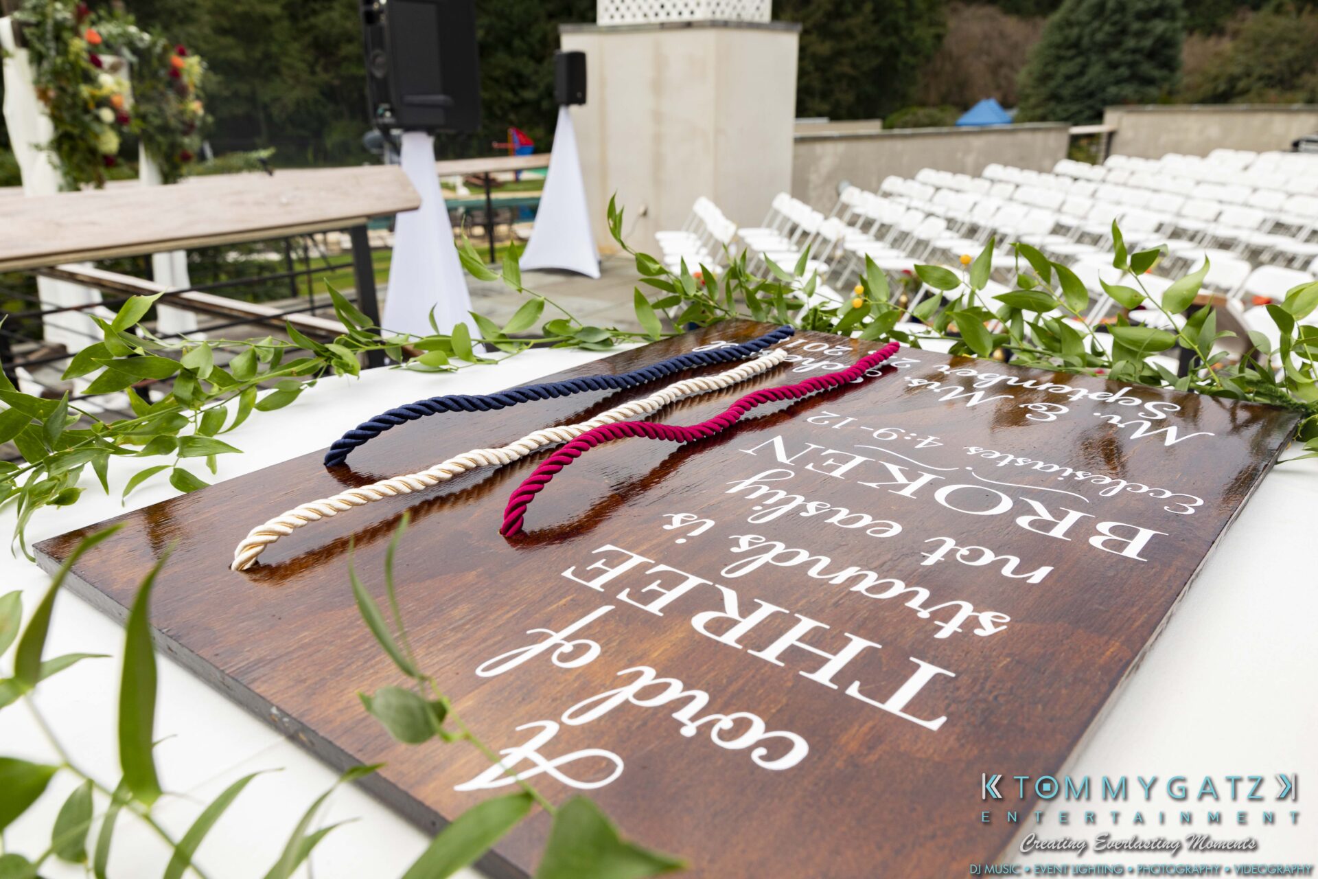 Lisa-Robert-092218-BLOG-selections-WM-39 - TOMMY GATZ ENTERTAINMENT Wedding altar table with three colored cords on a wooden sign displaying a quote. White chairs and outdoor setting in the background.