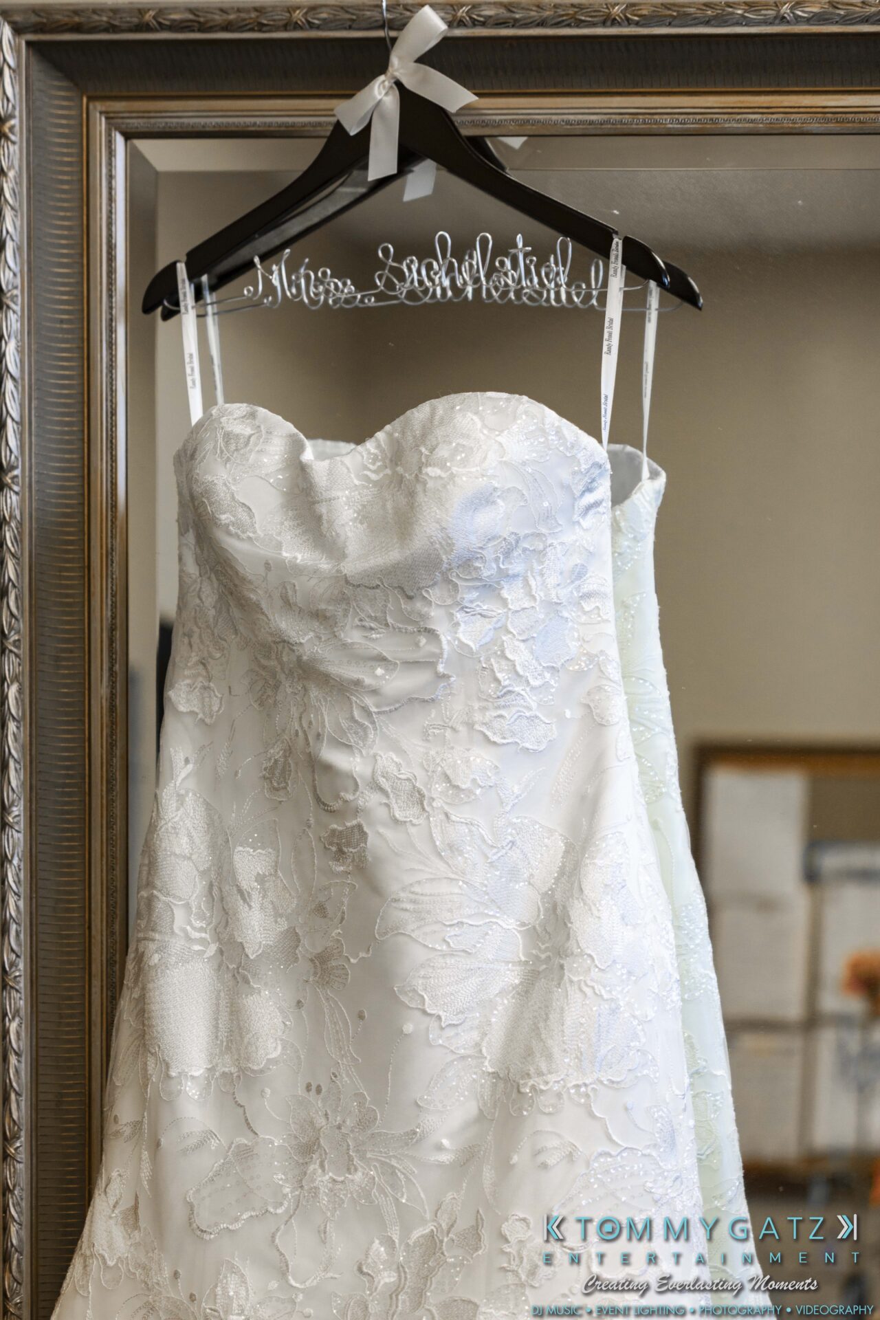 Lisa-Robert-092218-BLOG-selections-WM-4 - TOMMY GATZ ENTERTAINMENT A white lace wedding dress hangs on a personalized hanger in front of a mirror with a decorative frame.