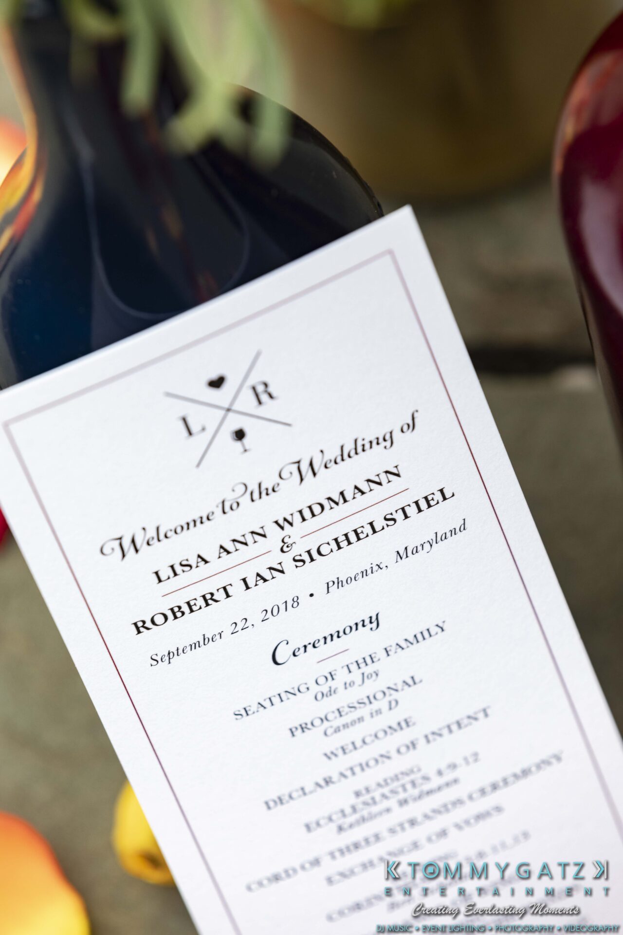 Lisa-Robert-092218-BLOG-selections-WM-41 - TOMMY GATZ ENTERTAINMENT Close-up of a wedding program featuring the names Lisa Ann Widmann and Robert Ian Sichelstiel, dated September 22, 2018, in Phoenix, Maryland, placed against a wine bottle.