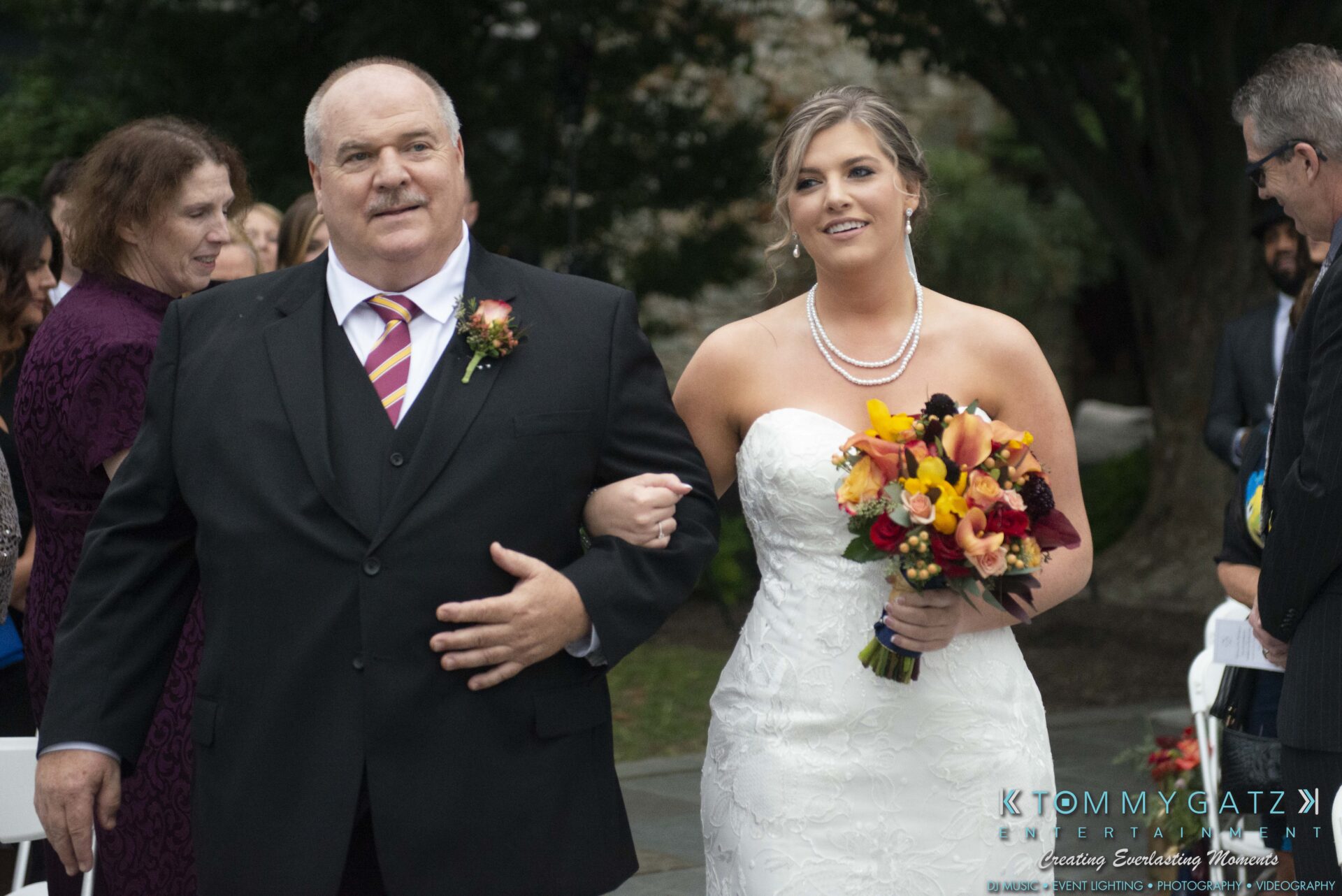 Lisa-Robert-092218-BLOG-selections-WM-50 - TOMMY GATZ ENTERTAINMENT A bride in a white dress and pearl necklace walks down an outdoor aisle with an older man in a suit. She holds a bouquet of colorful flowers.
