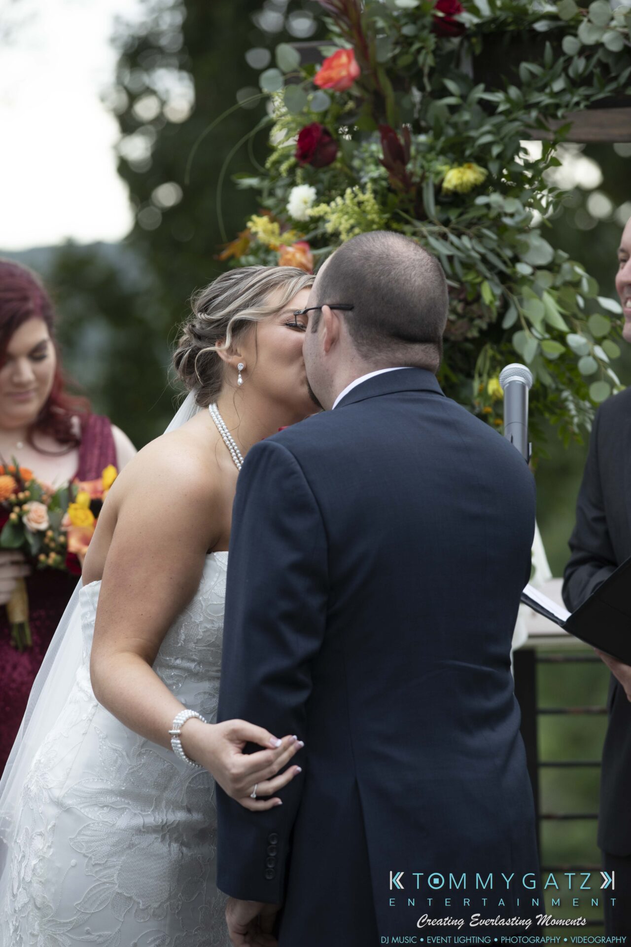 Lisa-Robert-092218-BLOG-selections-WM-57 - TOMMY GATZ ENTERTAINMENT A bride and groom share a kiss during their wedding ceremony, with a partially visible officiant in the background holding flowers.
