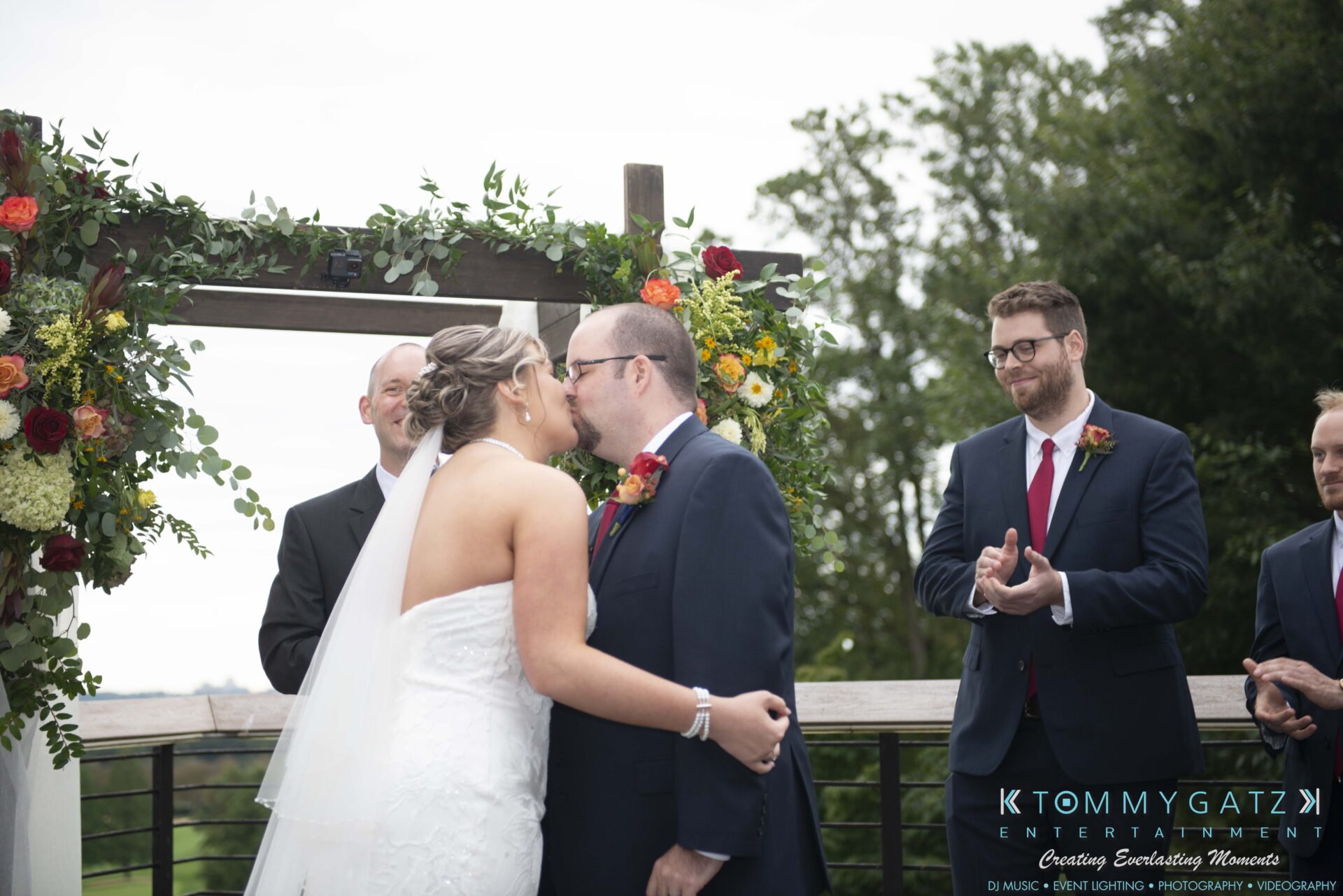 Lisa-Robert-092218-BLOG-selections-WM-58 - TOMMY GATZ ENTERTAINMENT A bride and groom kiss at an outdoor wedding ceremony, with attendants clapping. Floral arch and greenery are visible.